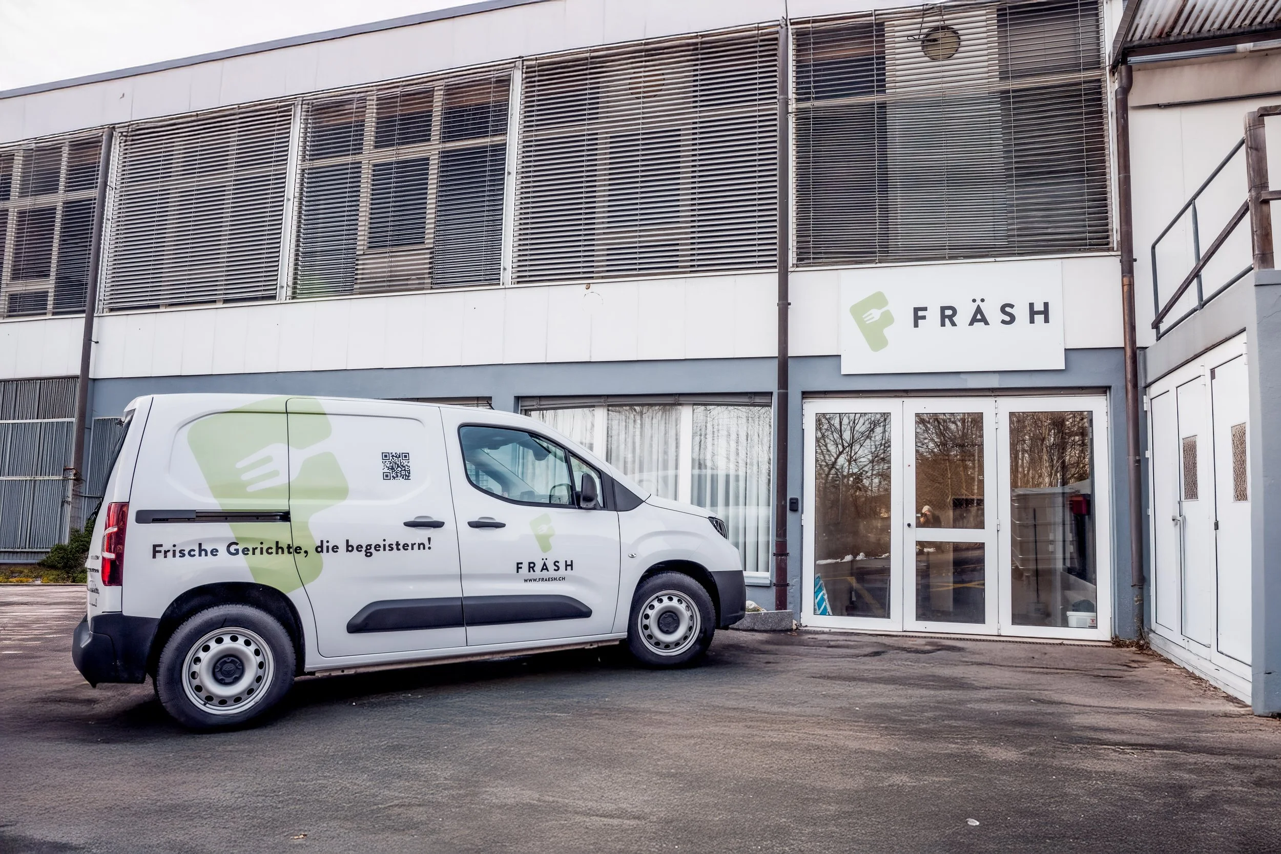 Vorne ist ein weißes Fahrzeug mit Werbung für Fräsh, einem Lieferservice für frische Gerichte, mit dem Slogan 'Frische Gerichte, die begeistern!' und einer QR-Code. Das Fahrzeug steht vor einem Gebäude mit einem Fräsh-Logo über den Eingangstüren.