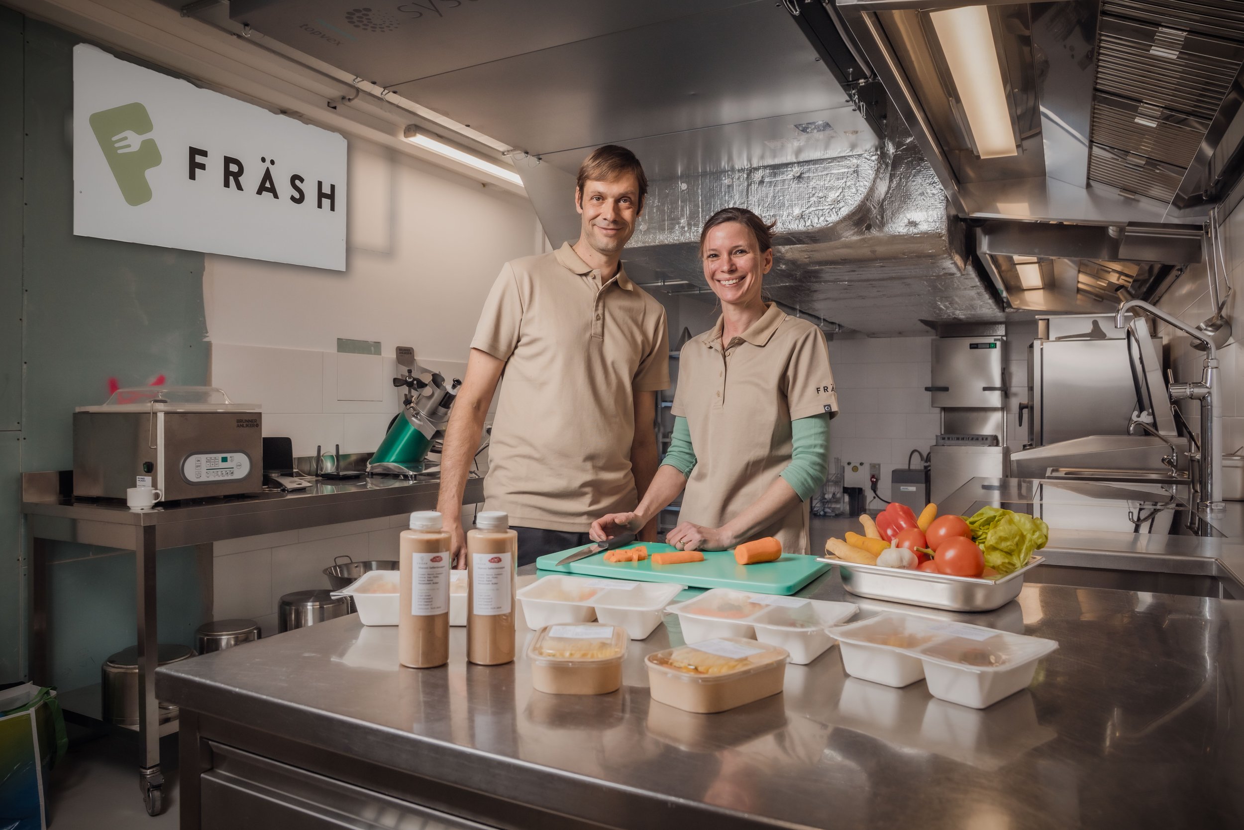 Zwei Menschen in Kochschürzen stehen in einer Küche, schneiden Gemüse und bereiten Mahlzeiten vor, im Hintergrund steht ein Schild mit der Aufschrift 'FRÄSH'.