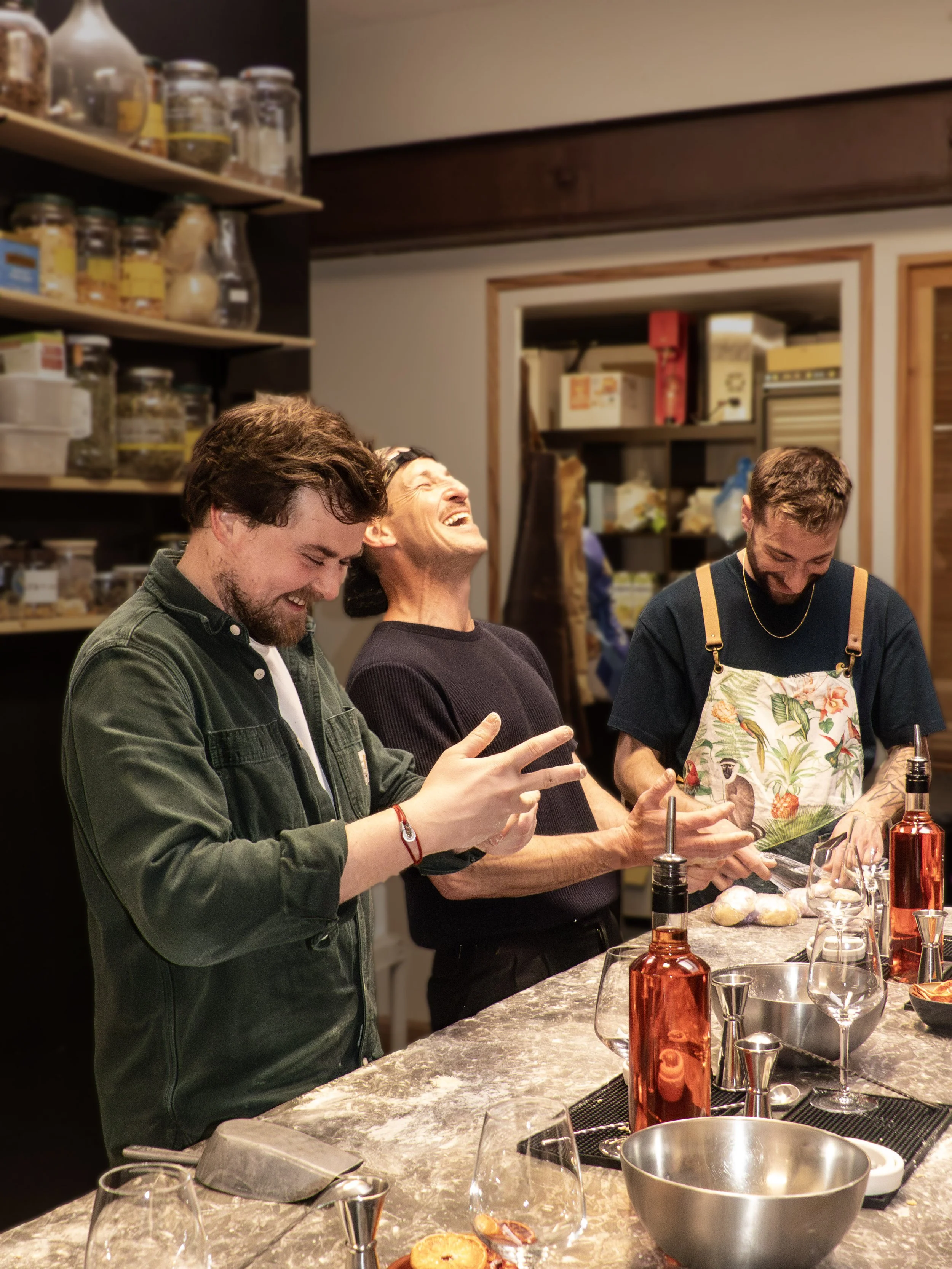 Trois hommes rient et sourient dans une cuisine moderne avec une étagère de bocaux en arrière-plan, en préparant des ingrédients sur un comptoir en marbre.