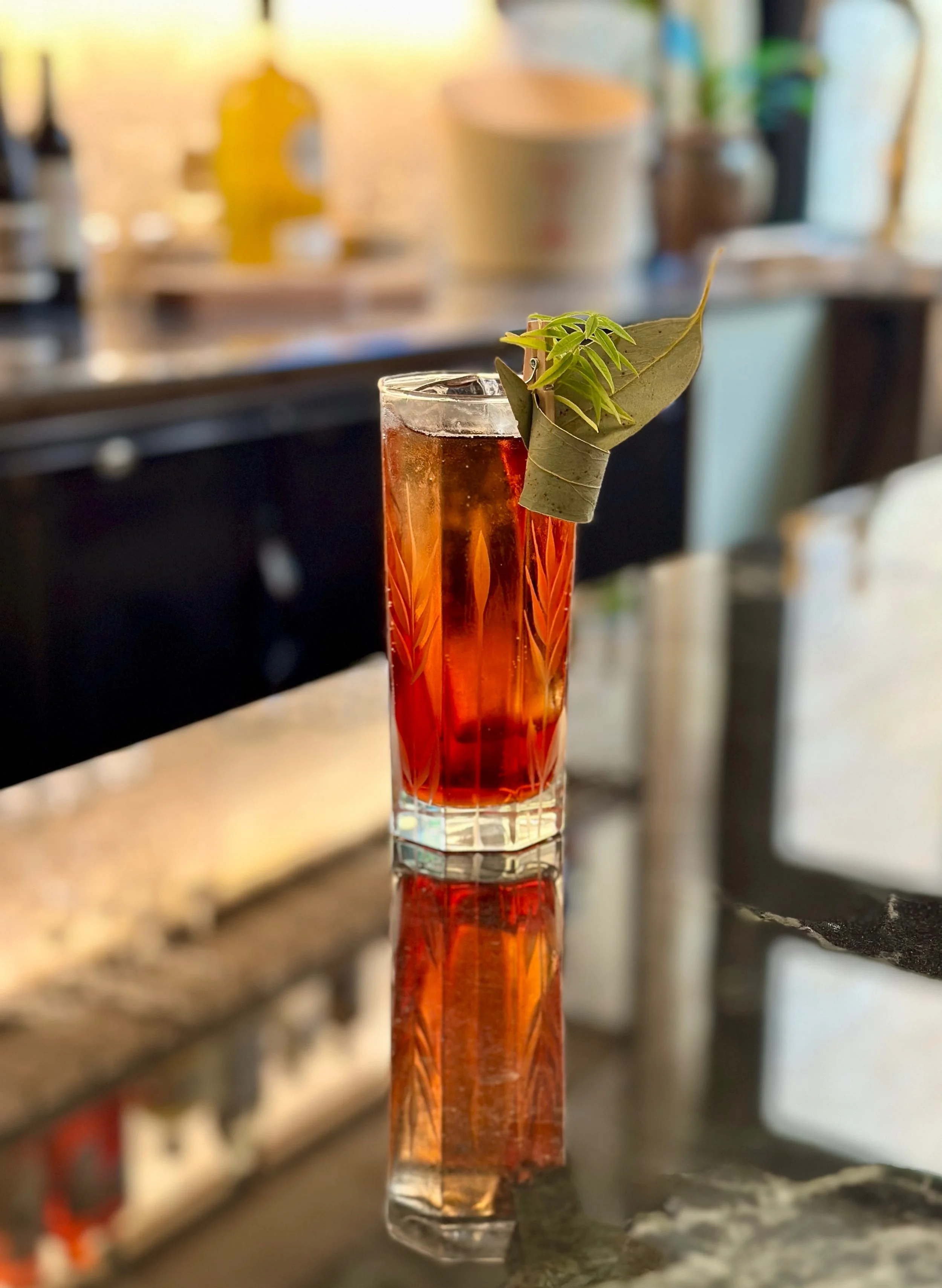 Un cocktail dans un verre tall, décoré d'une feuille de menthe et d'une feuille d'eucalyptus. La boisson est rougeâtre avec des glaçons, posé sur un comptoir en marbre.