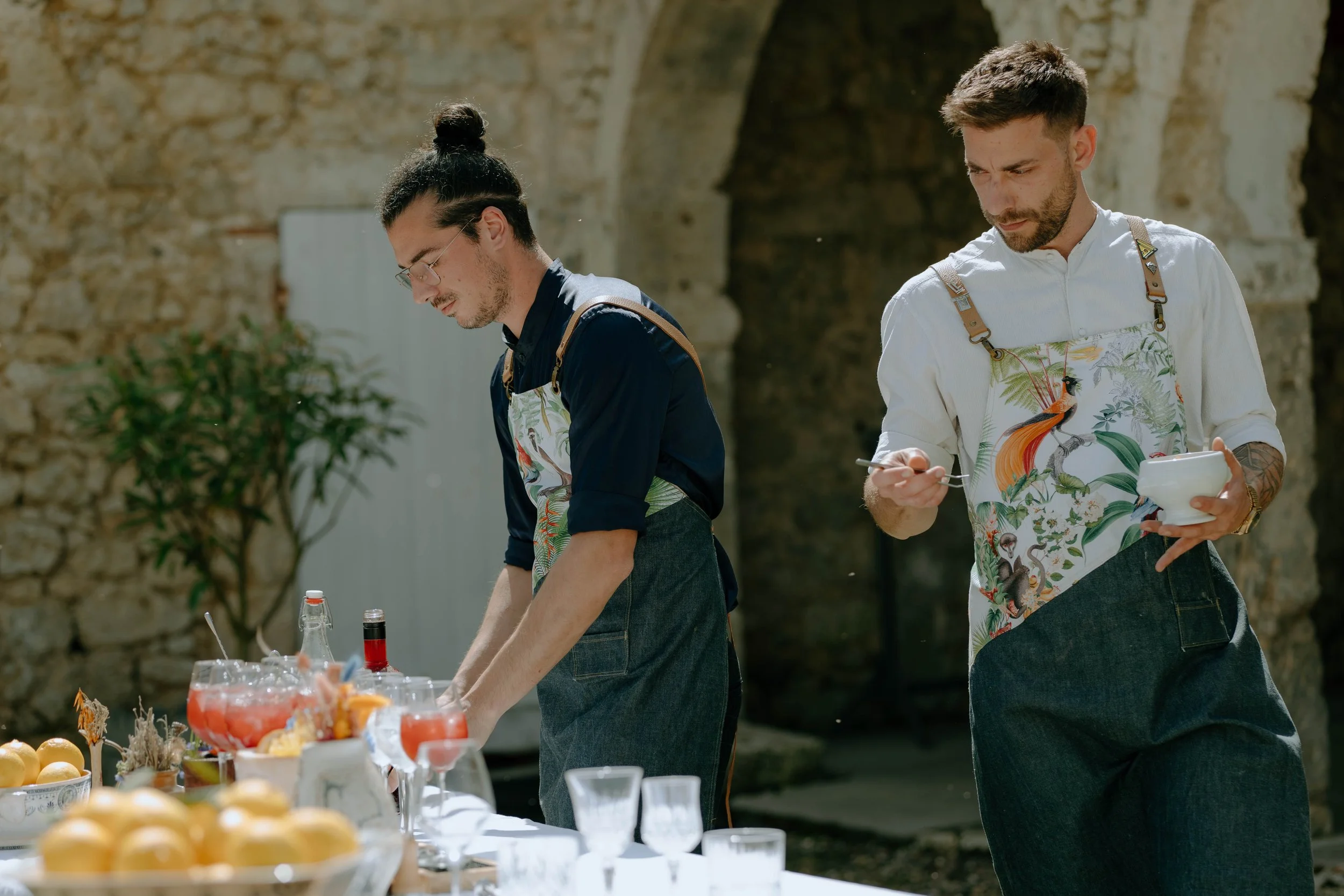 Deux hommes préparent des boissons lors d'une fête en extérieur, portant des tabliers avec un motif floral tropical, devant une table avec des verres et des fruits.