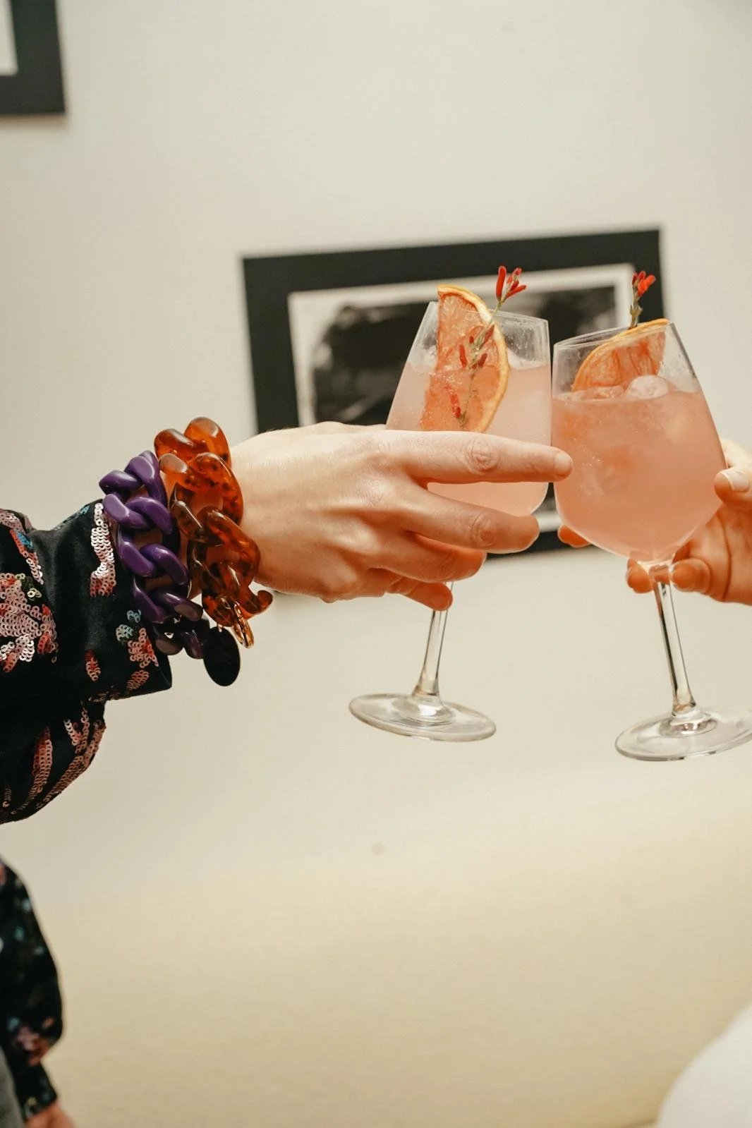 Deux personnes trinquent avec des verres de cocktail rose, garnis de tranche d'orange et de petites brochettes de cerise. Une personne porte des bracelets en plastique violet et orange.