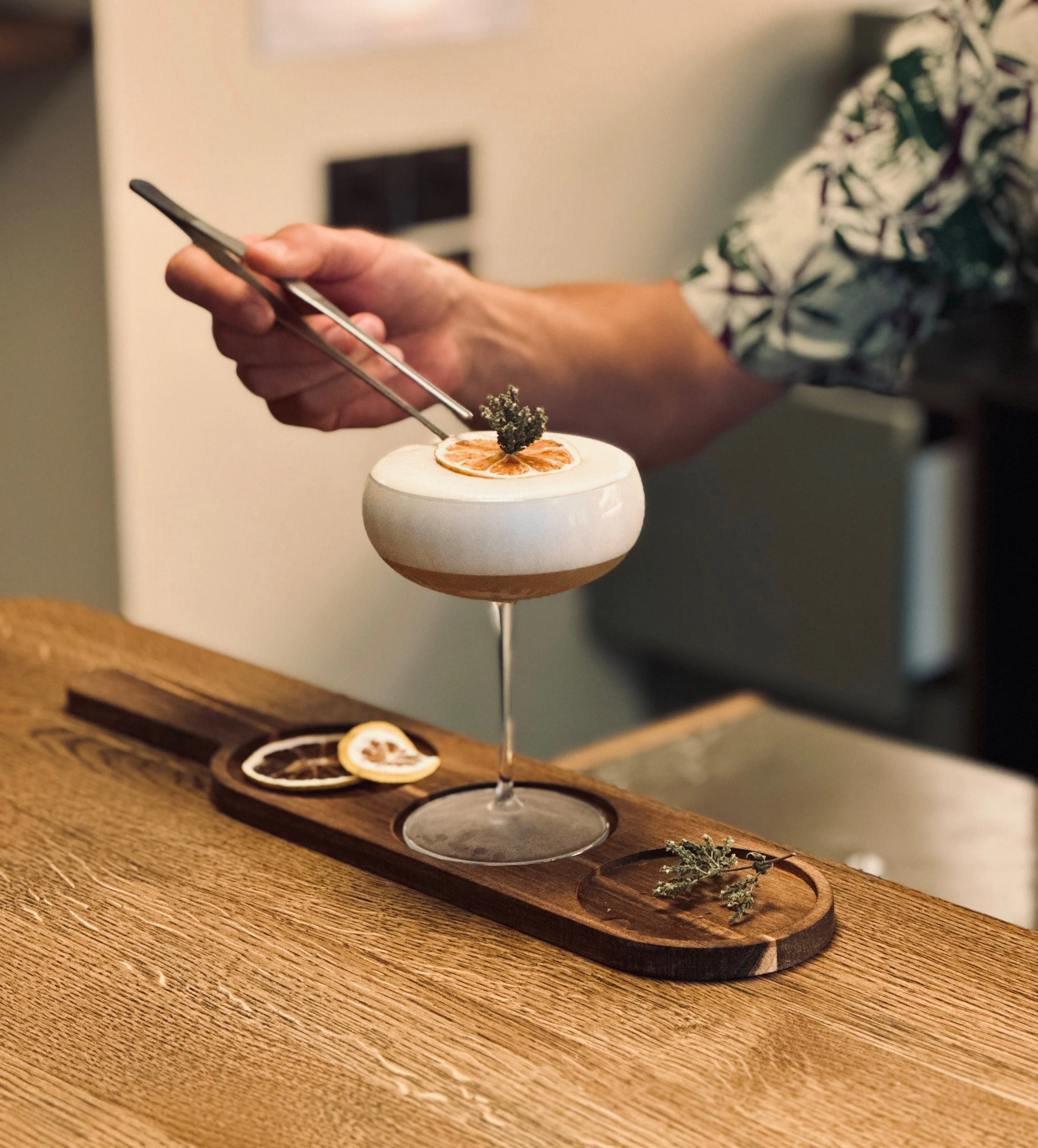 Un cocktail dans un verre à pied avec des décorations d'agrumes et de plantes, posé sur un plateau en bois sur une table en bois.