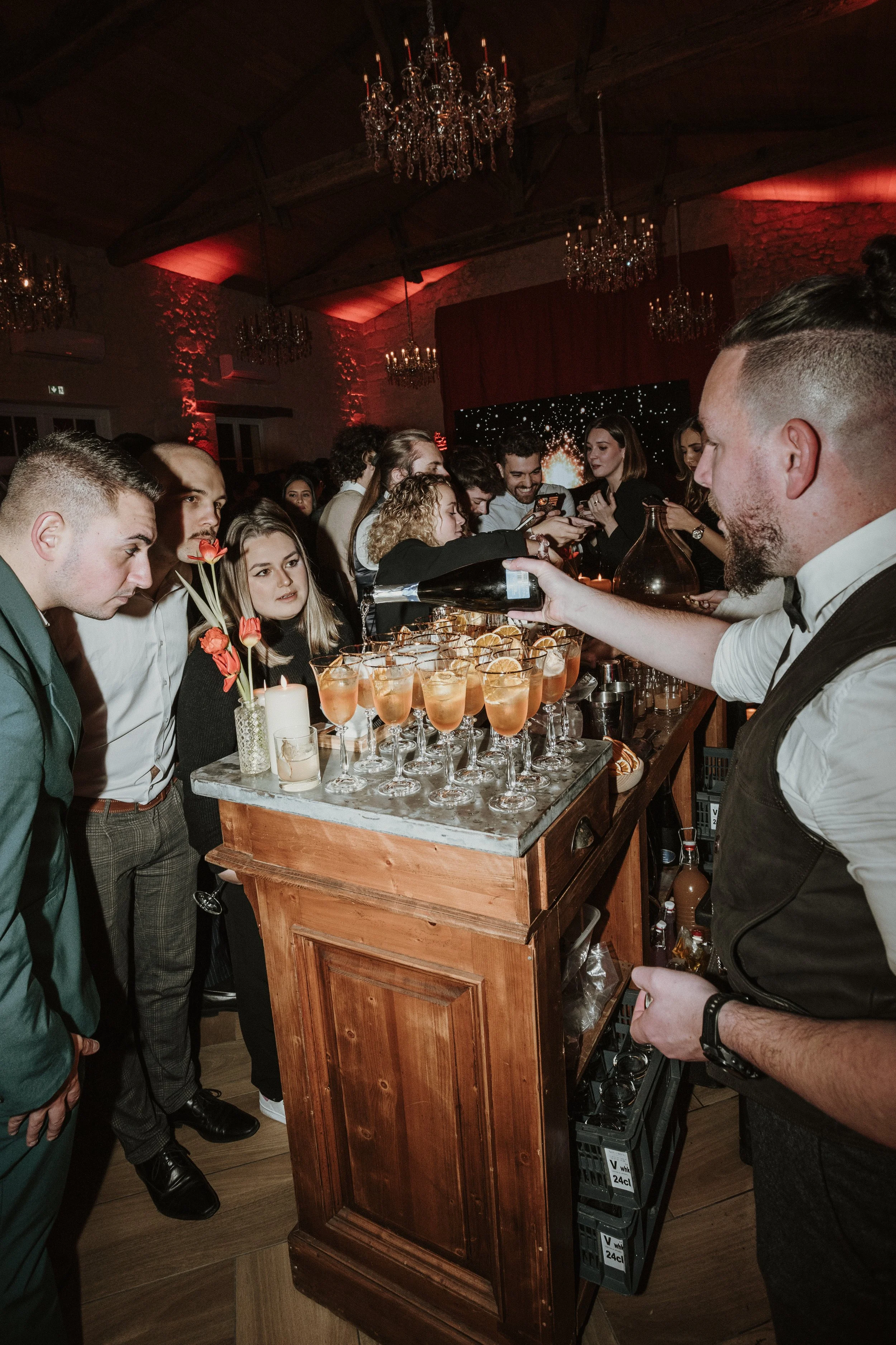 Un groupe de personnes boit des cocktails au bar lors d'une soirée dans une salle élégante avec chandeliers et éclairage tamisé.