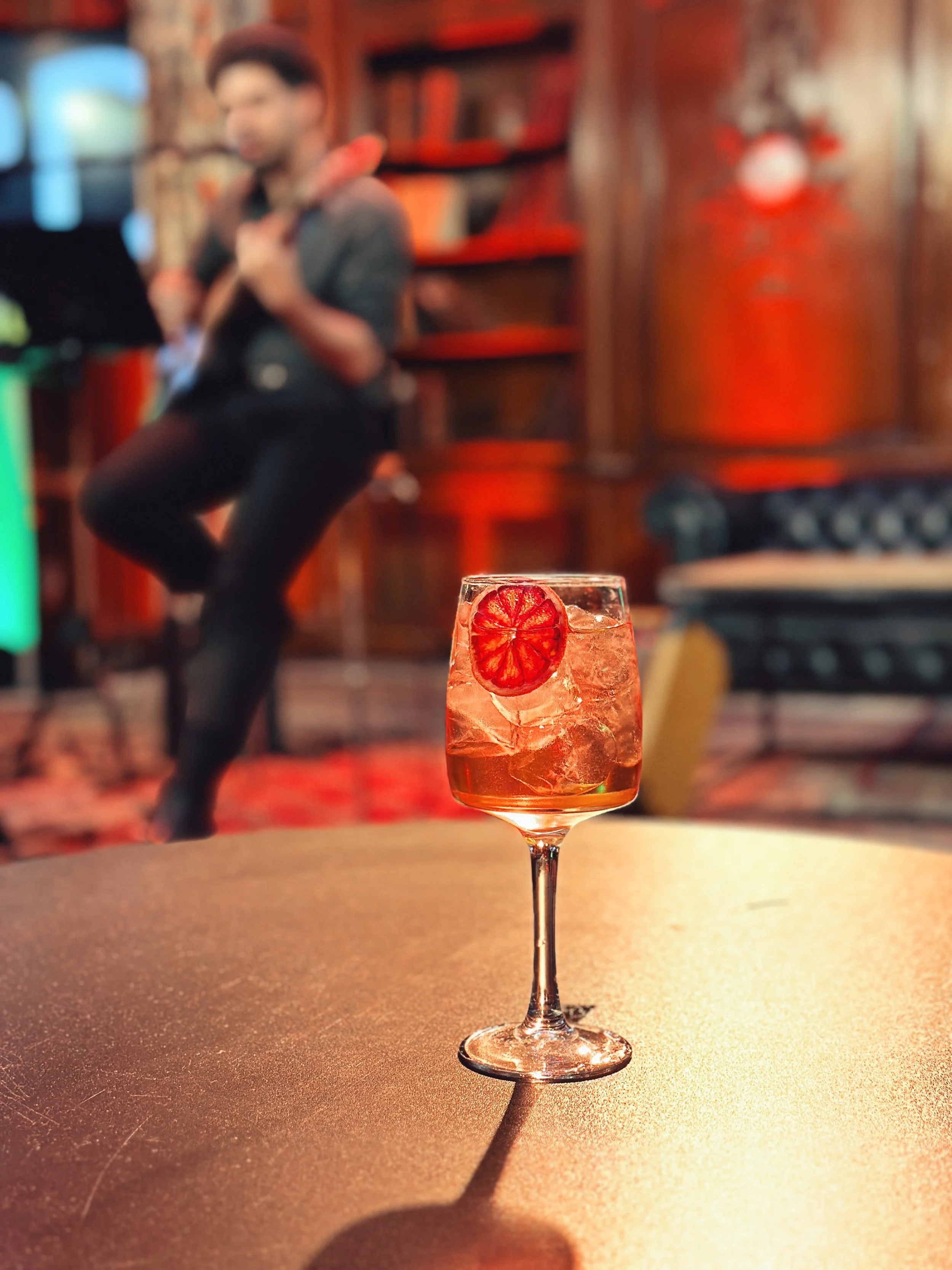 Sunset Spirit 2025, un Spritz placé sur une table en première plan, avec un musicien qui joue de la guitare dans un fond flou.