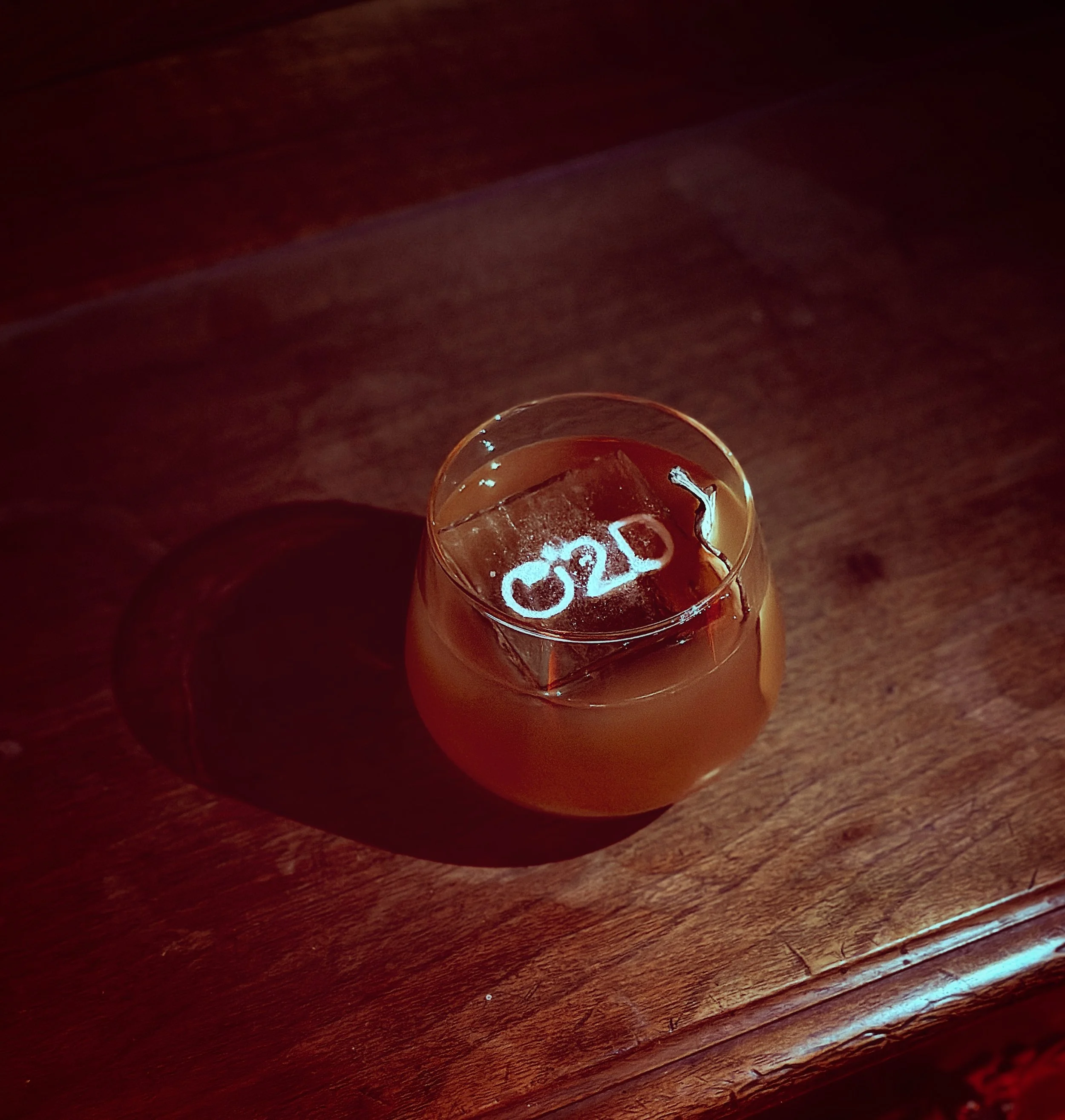 Un verre de cocktail avec un gros glaçon sur une table en bois, la glace porte une inscription 'O2D'.