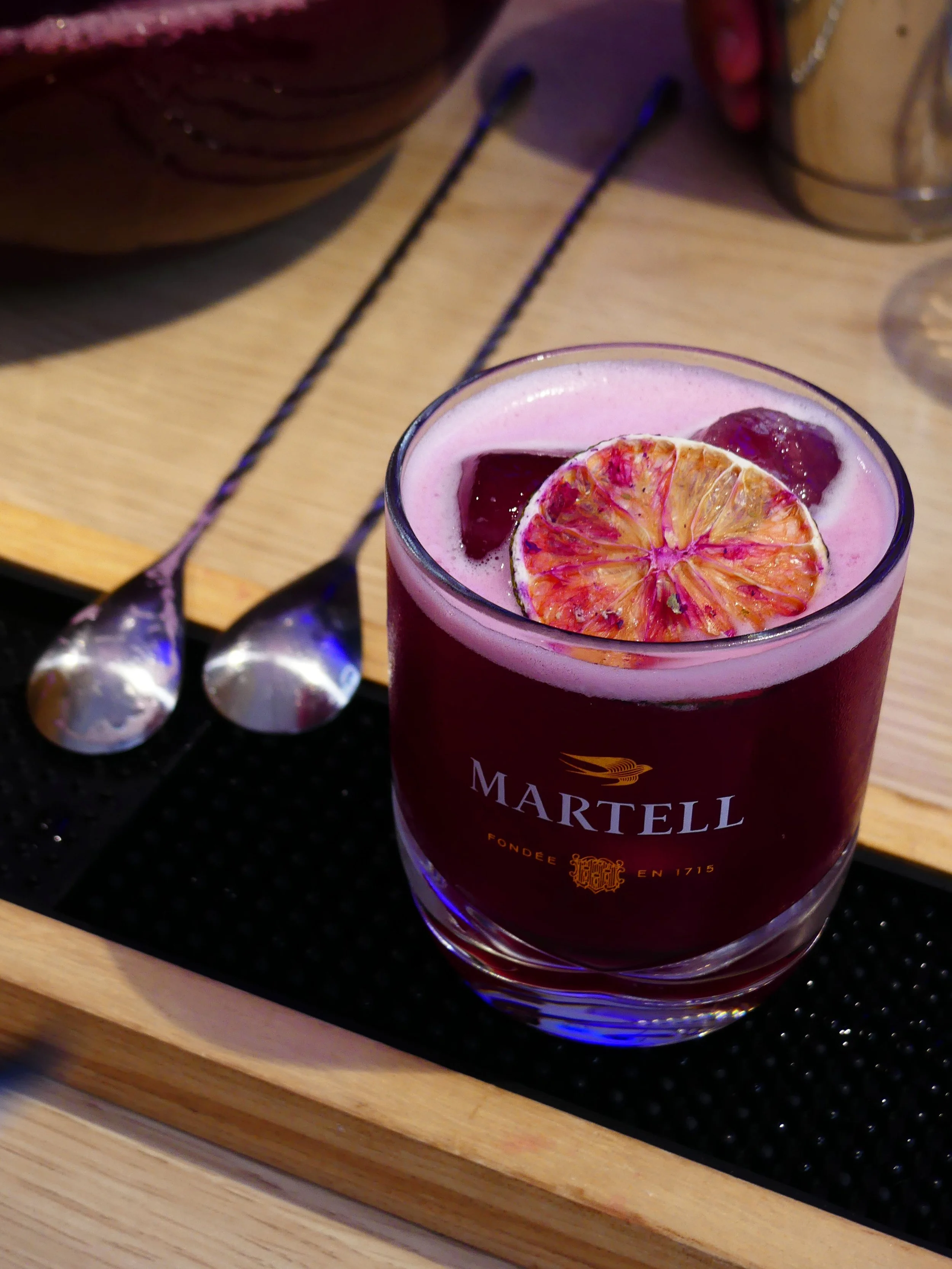 Sunset Spirit 2025, Verre de cocktail rouge avec une tranche de citron et des fruits rouges, posé sur un plateau en bois