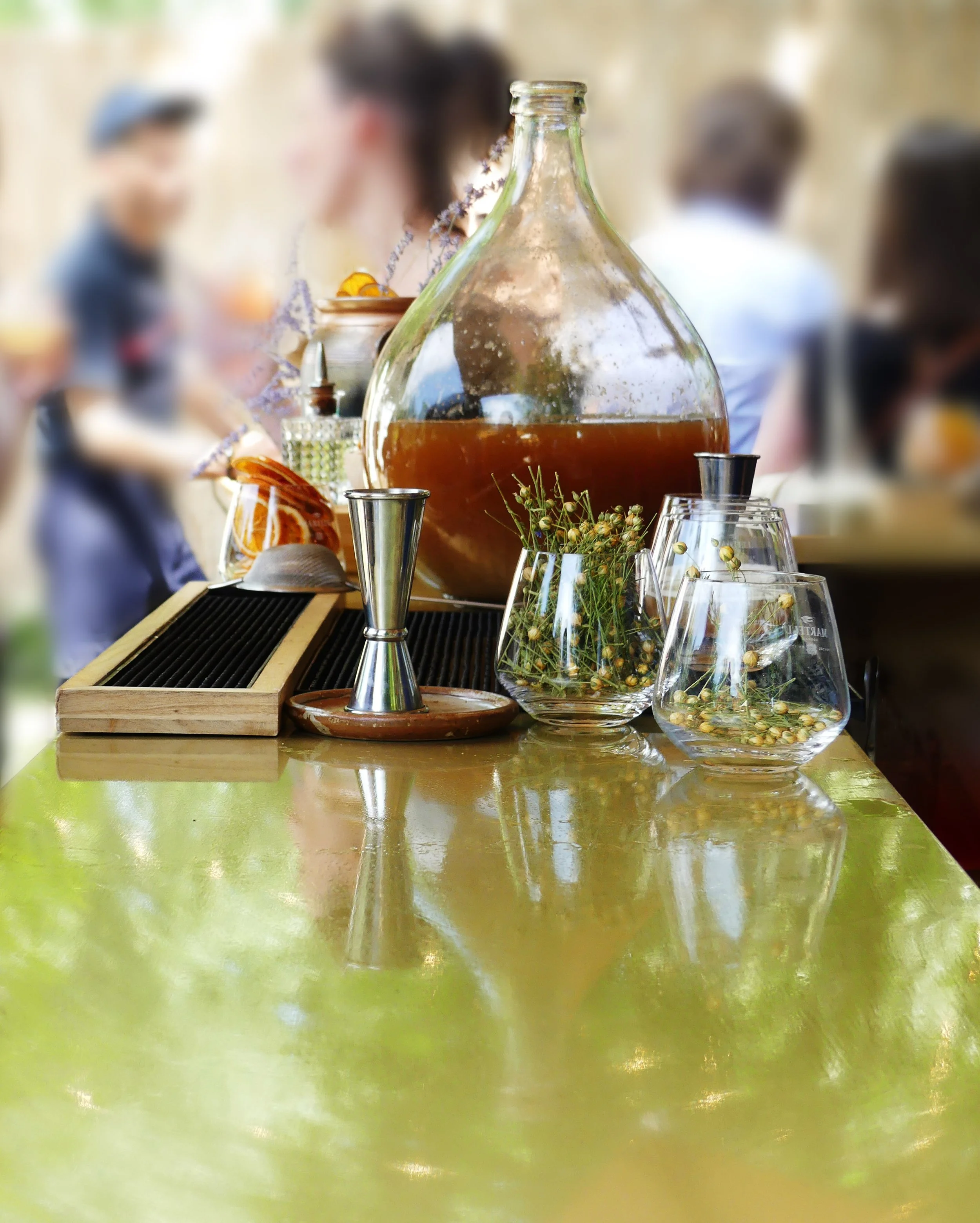 Une table en bois avec un grand bocal en verre rempli de boisson brune, entouré de verres avec des plantes séchées, un jigger en métal, un plateau en bois avec des accessoires de bar, et un décor flou avec plusieurs personnes en arrière-plan dans un cadre intérieur lumineux.