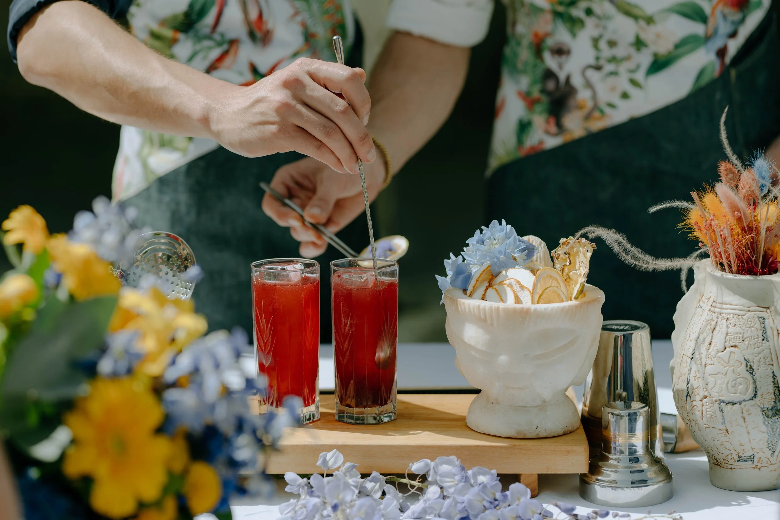 Deux cocktails rouges dans des verres hauts, décorés avec des pailles métalliques, entourés de fleurs colorées et de vaisselle élégante sur une table en extérieur.