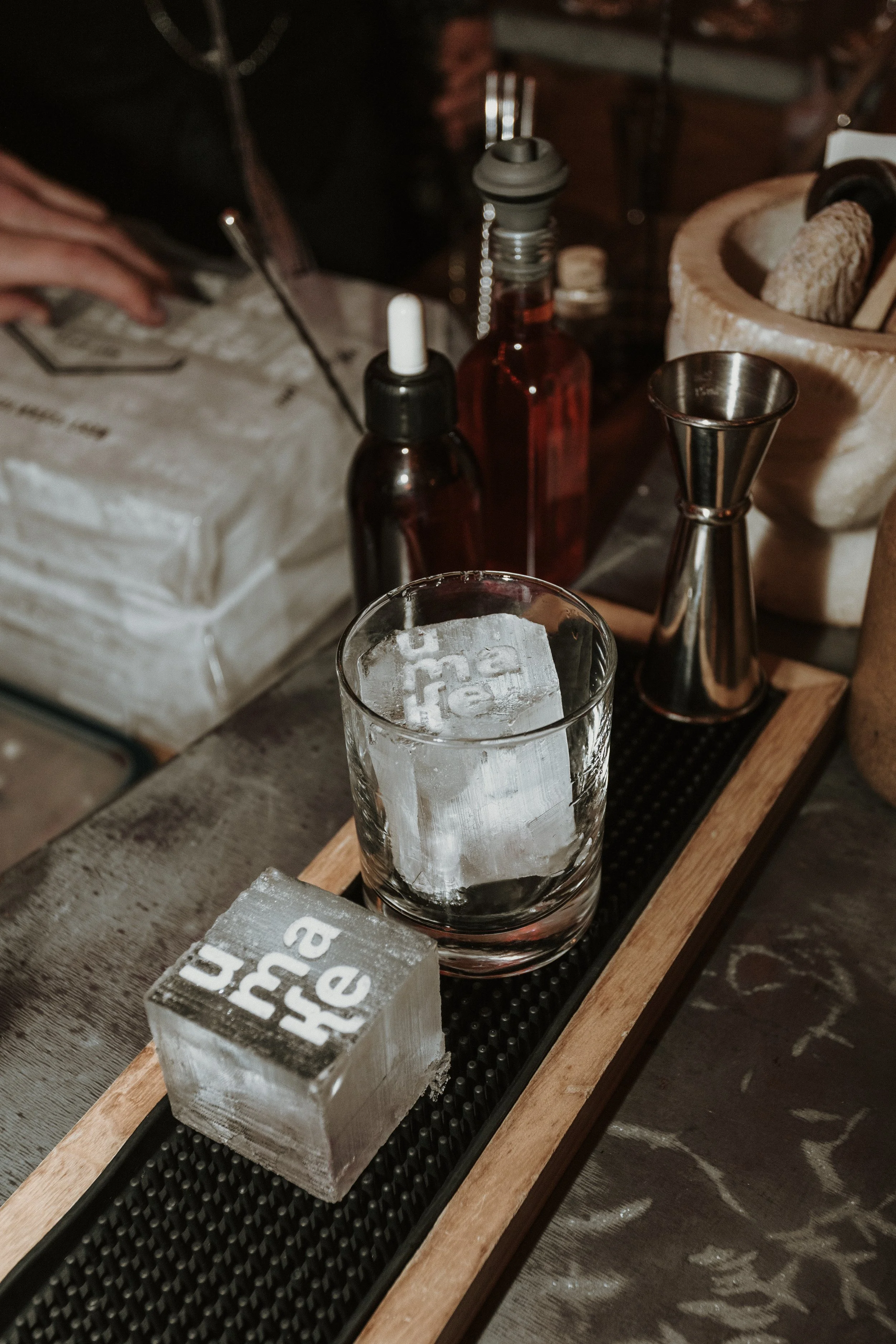 Sunset Spirit 2025, Verre de cocktail avec un gros glaçon sur un plateau en bois, avec un autre glaçon et un jigger à côté, dans un bar.