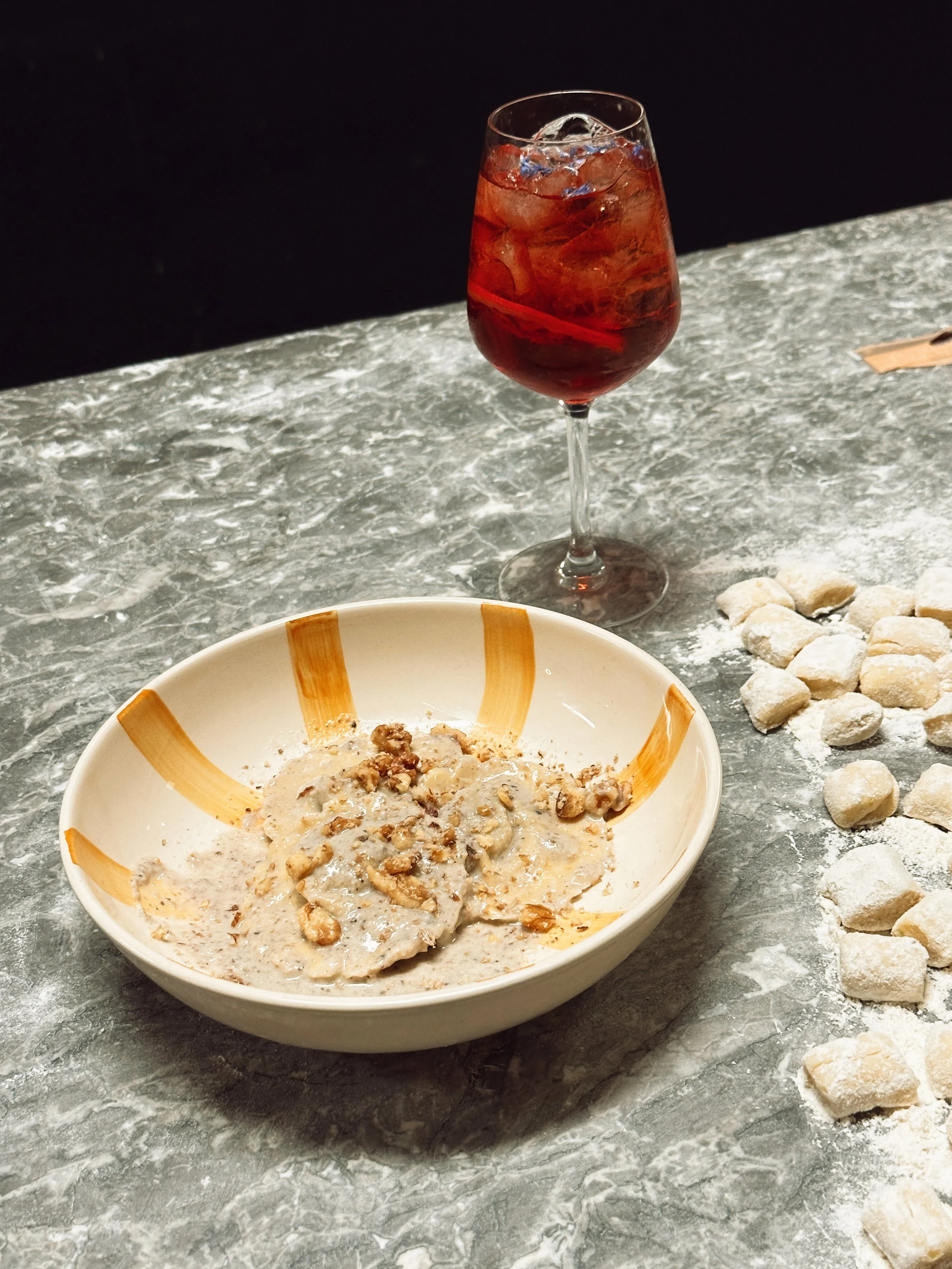 Un verre de vin rouge avec glace, un bol de gnocchis à la sauge et à la crème, et des morceaux de gnocchis recouverts de farine sur une table en marbre.