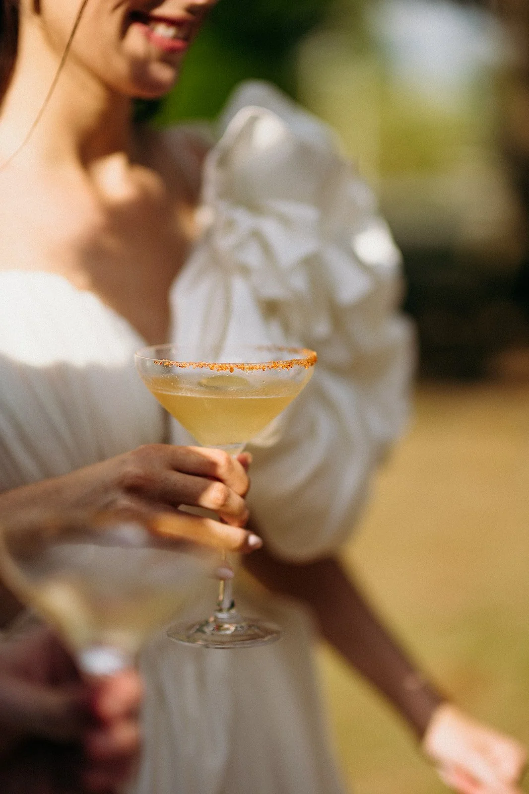 Une femme en robe blanche tient un verre à cocktail avec une boisson jaune et une touche de piment en bordure, lors d'une occasion festive extérieure.