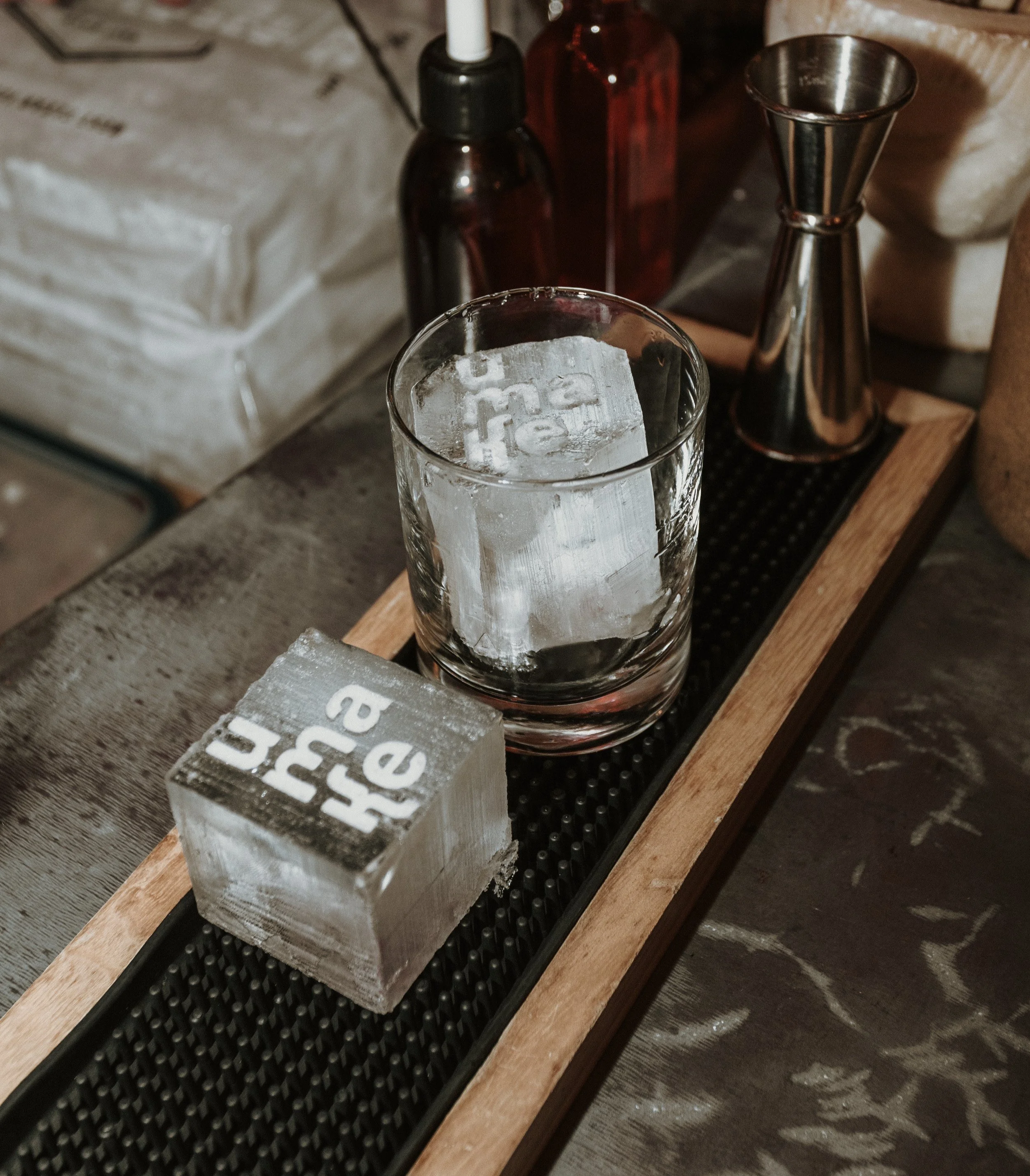 Verre et glaçons en forme de cube avec inscriptions, sur un plateau en bois, avec divers bouteilles et ustensiles en arrière-plan.