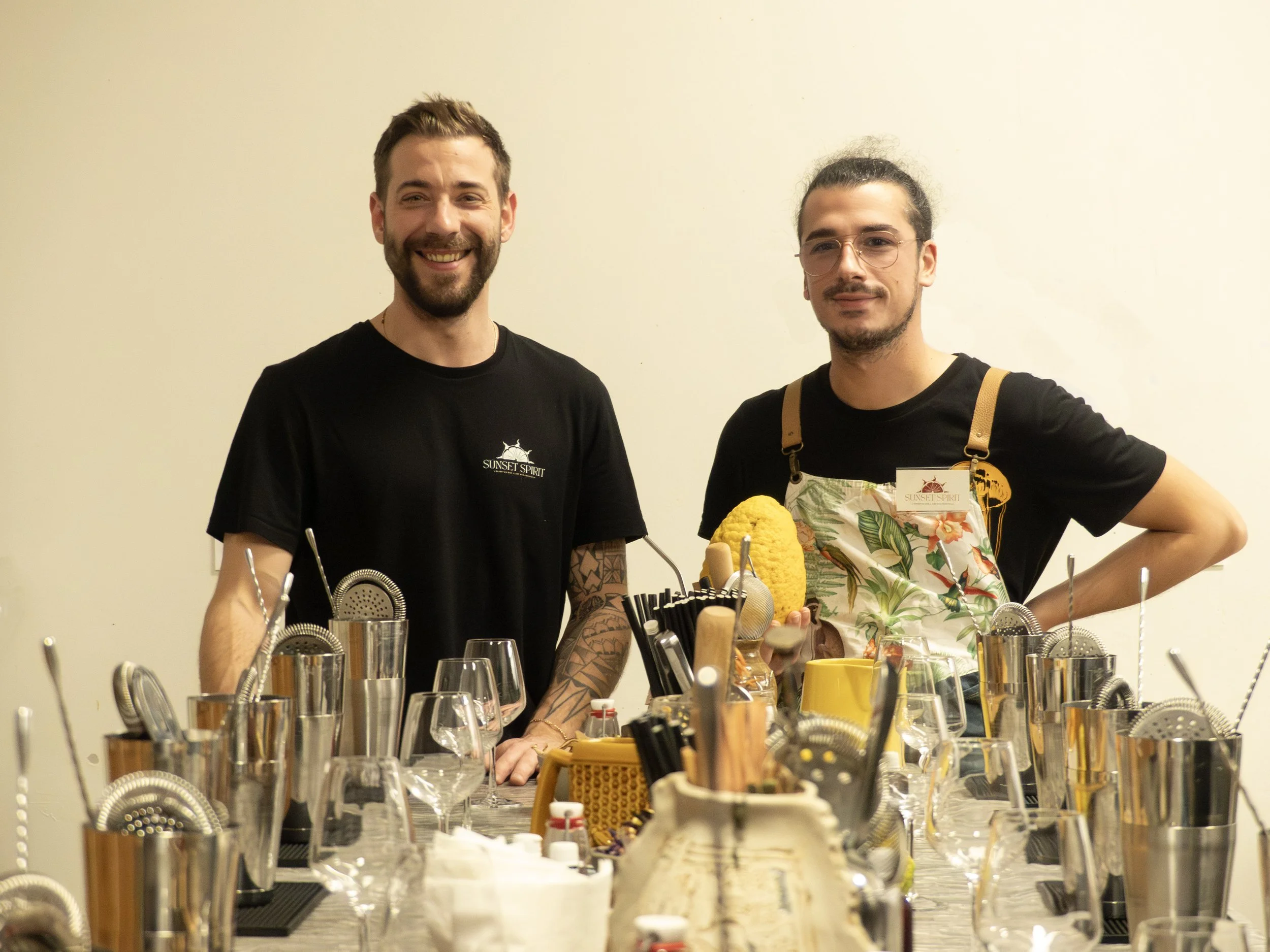 Deux hommes debout derrière une table de cuisine, entourée d'ustensiles et de verres à vin, dans une pièce à mur blanc.