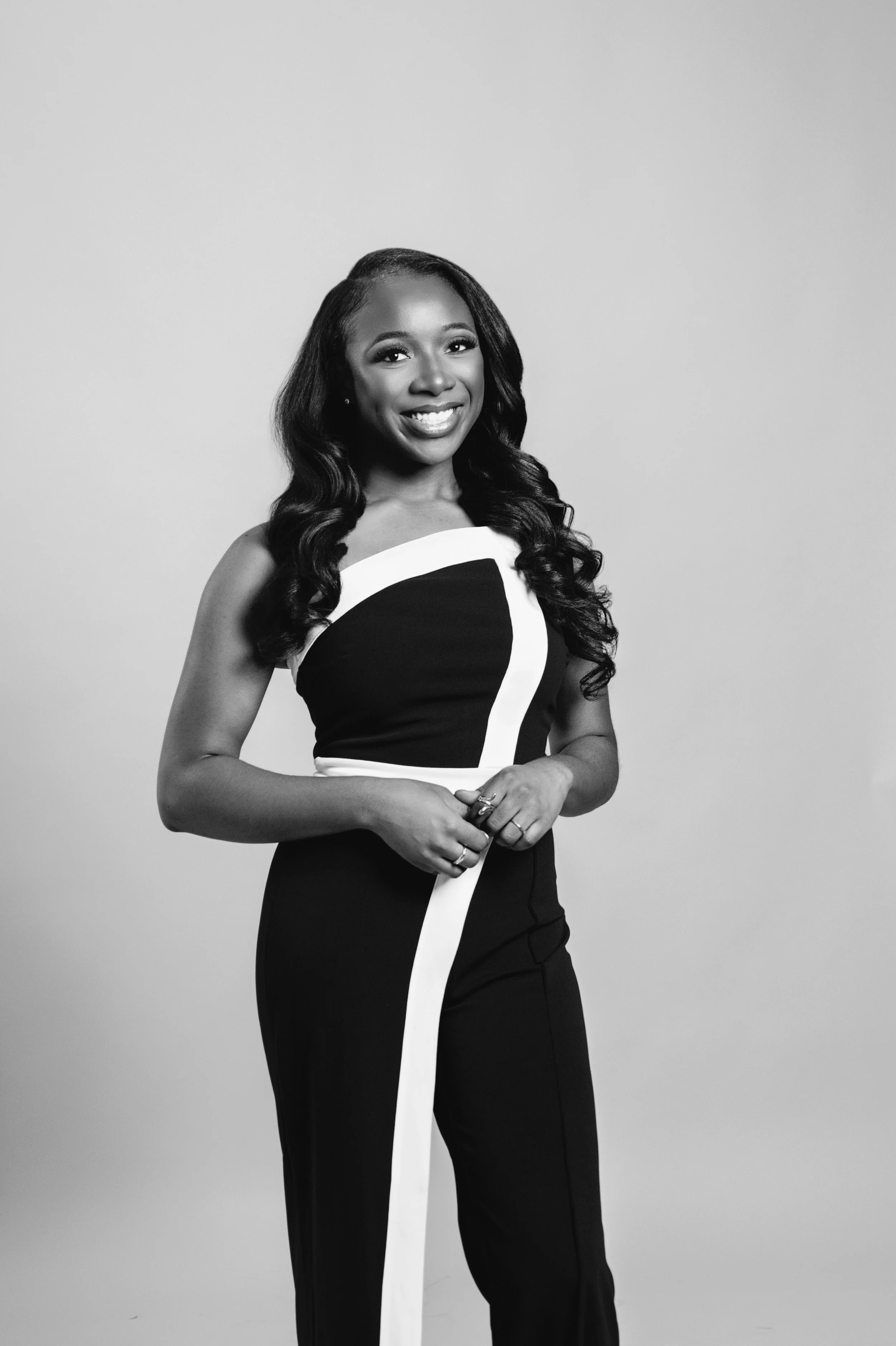 Black and white portrait of a woman smiling, wearing a sleeveless fitted dress with a bold white stripe design, standing against a plain background.