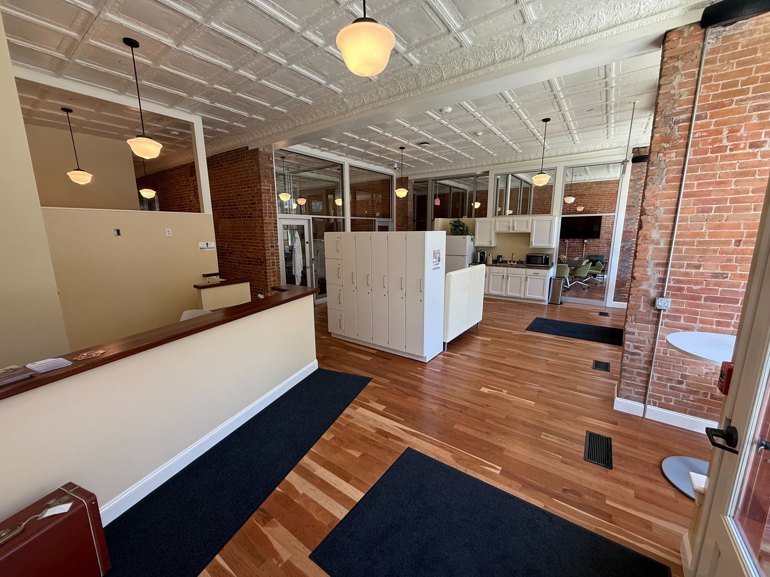 Interior of a modern office space with wooden flooring, brick walls, and pendant lighting. Features include a reception desk, lockers, a kitchenette, and conference room with a TV. Natural light enters through large windows.