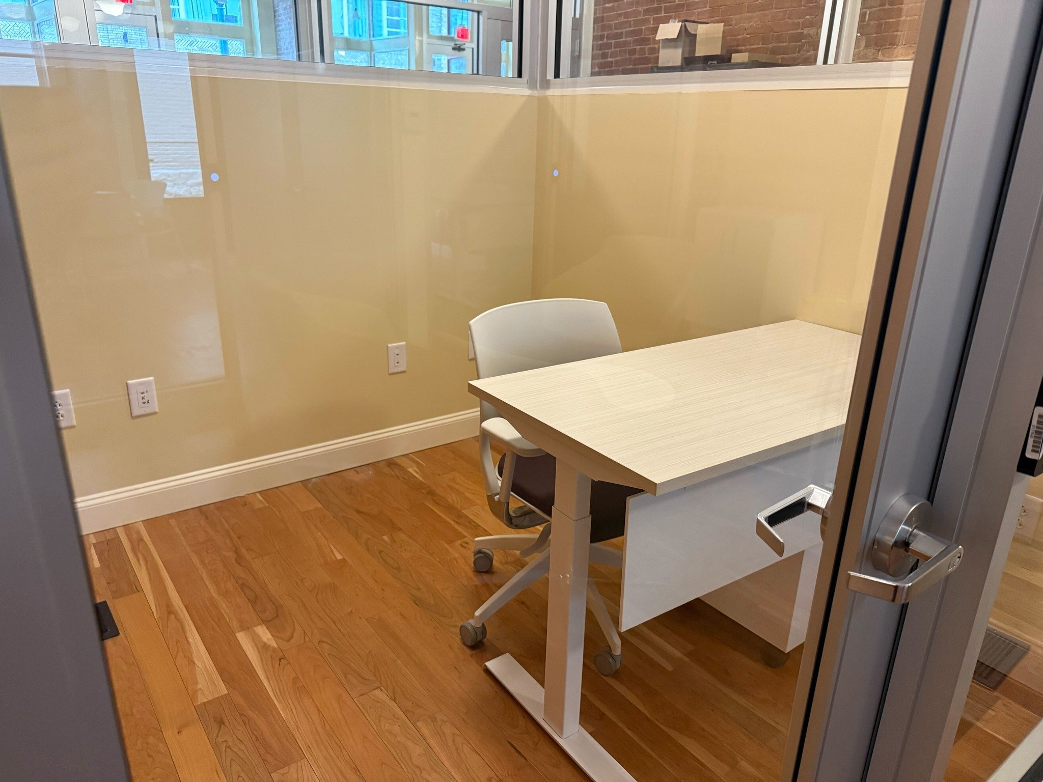 Compact office space with a wooden desk, a white chair on wheels, hardwood flooring, and glass walls.