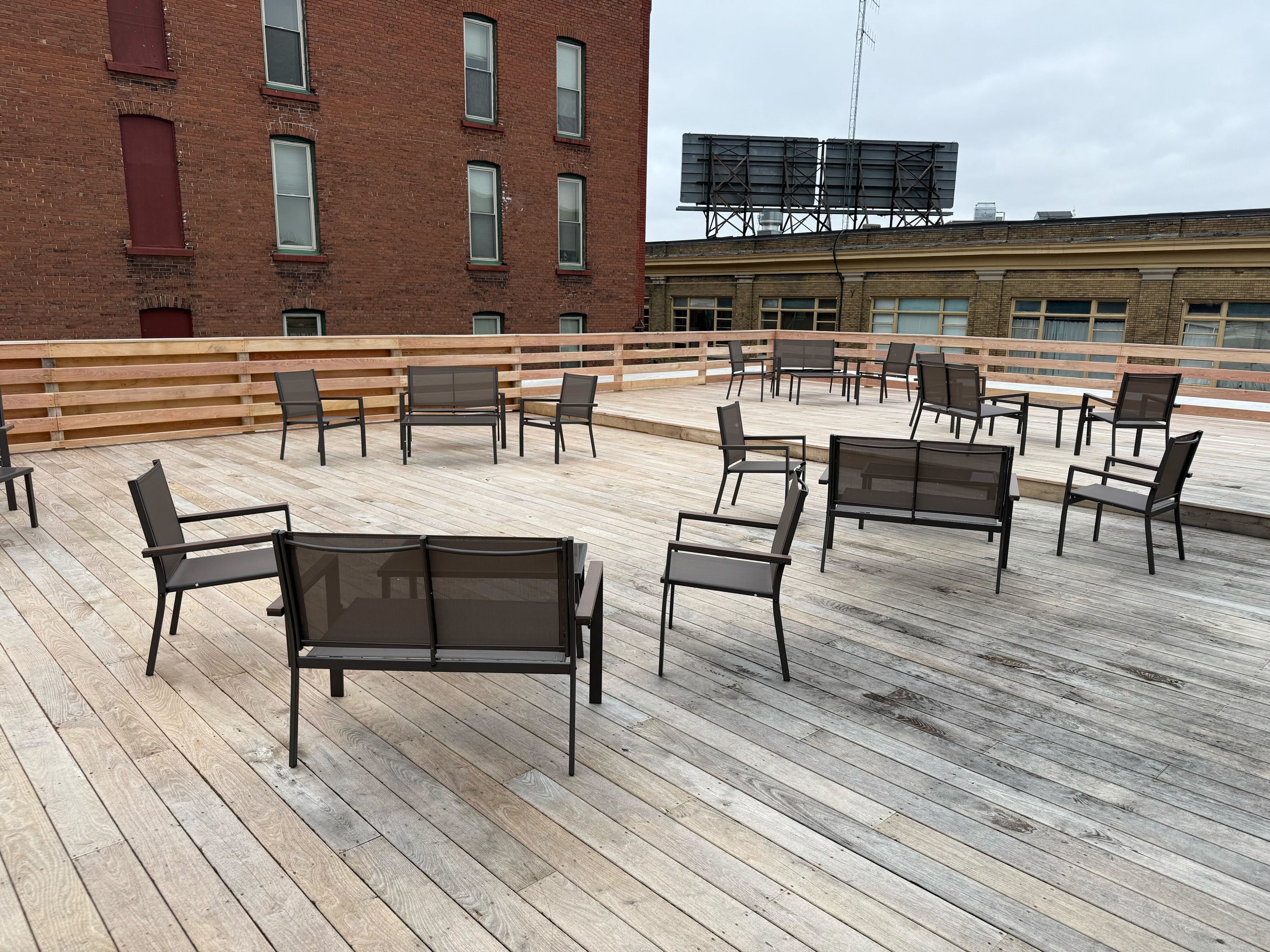 Outdoor wooden deck with several empty metal chairs and benches, surrounded by buildings and a large billboard in the background.