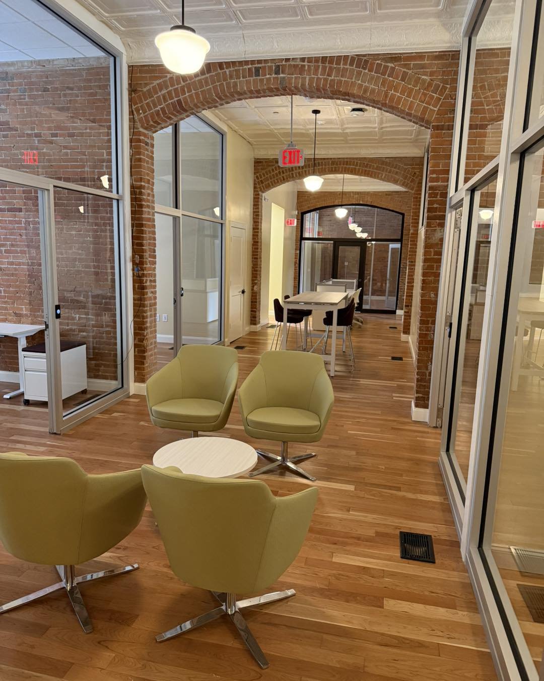 Modern office space with glass walls, brick arches, and wood flooring featuring green swivel chairs and tables.