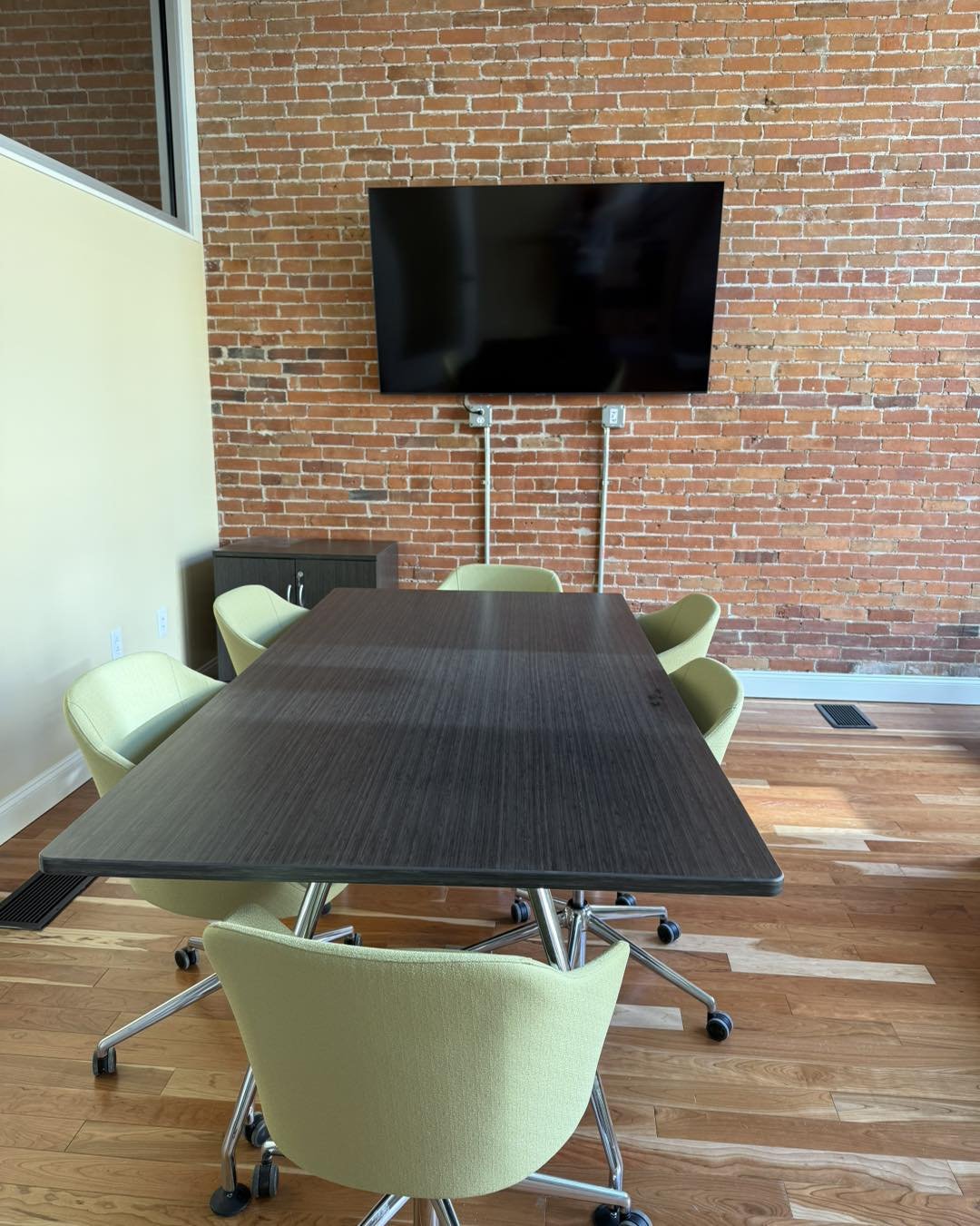 Modern conference room with a long table, green chairs, a large flat-screen TV on a brick wall, and wooden flooring. This is the small conference room in JCC's co-working space, the NEST.