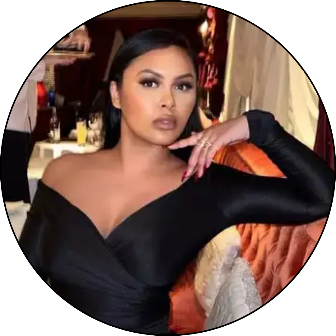 A woman with dark hair, makeup, and a black off-the-shoulder top, sitting on a couch in a room with a busy background.