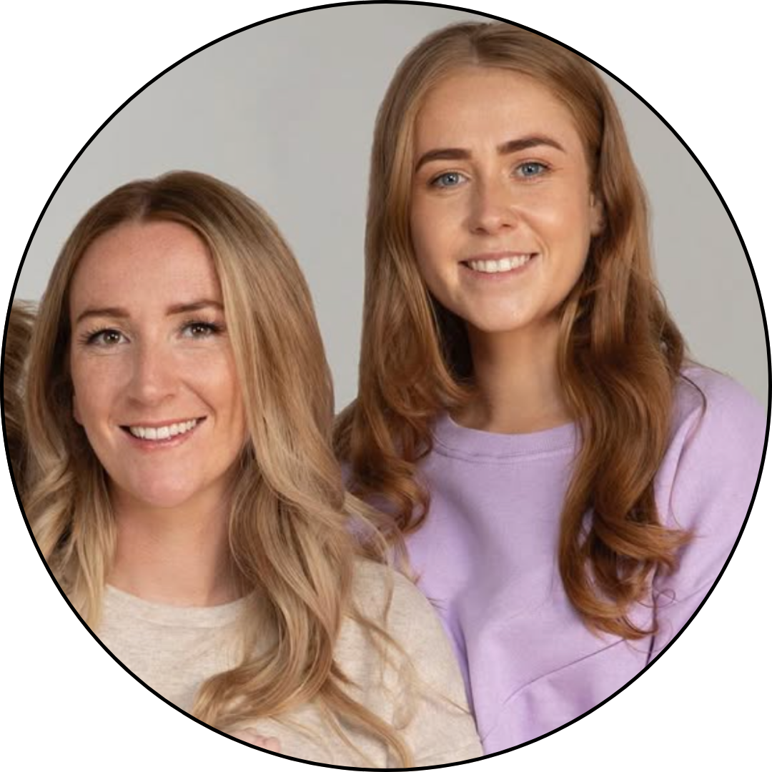 Two young women with light skin and long, wavy hair smiling, one has blonde hair and the other has red hair, wearing light-colored tops, against a neutral background.