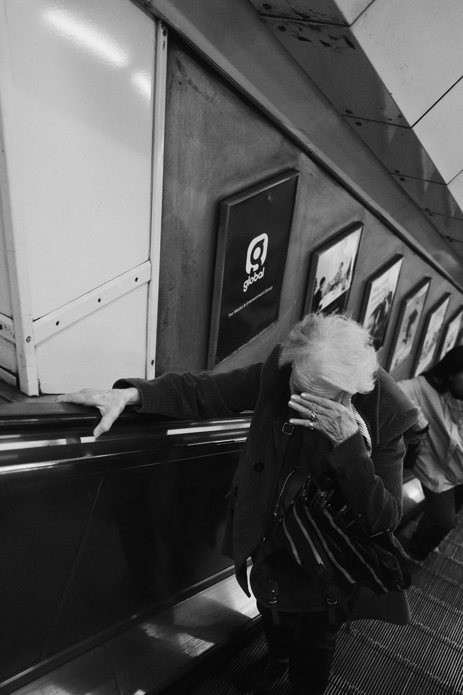 Une femme âgée en manteau sombre se couvre le visage avec une main dans un métro, soutenue par une bande d'acier. Elle porte des lunettes en bandoulière, et il y a un autre passager en arrière-plan. La photo est en noir et blanc, avec des affiches pu