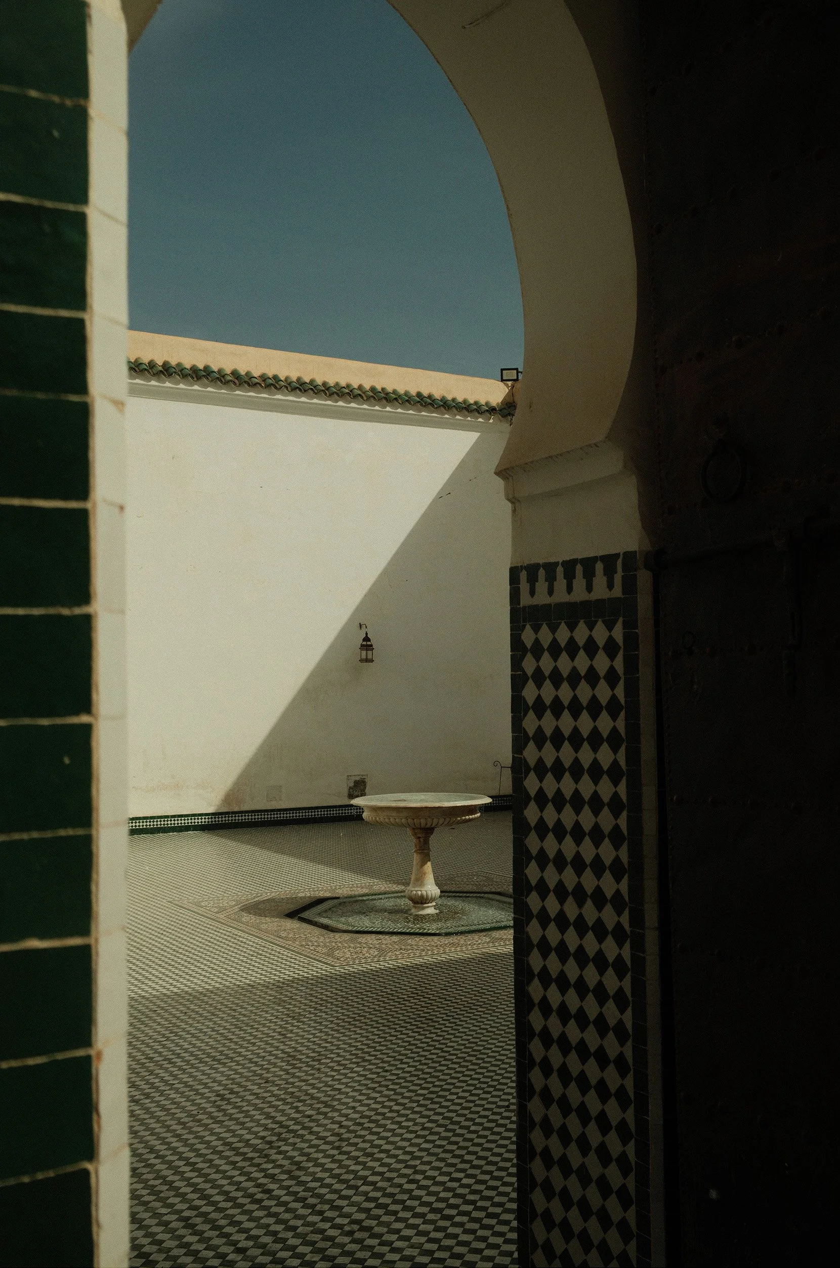 Vue d'une cour intérieure avec un sol en mosaïque, un mur blanc, une fontaine en pierre centrale, un mur orné de carreaux de motif géométrique et un ciel bleu en arrière-plan, vue partiellement encadrée par une arche et une porte en bois.