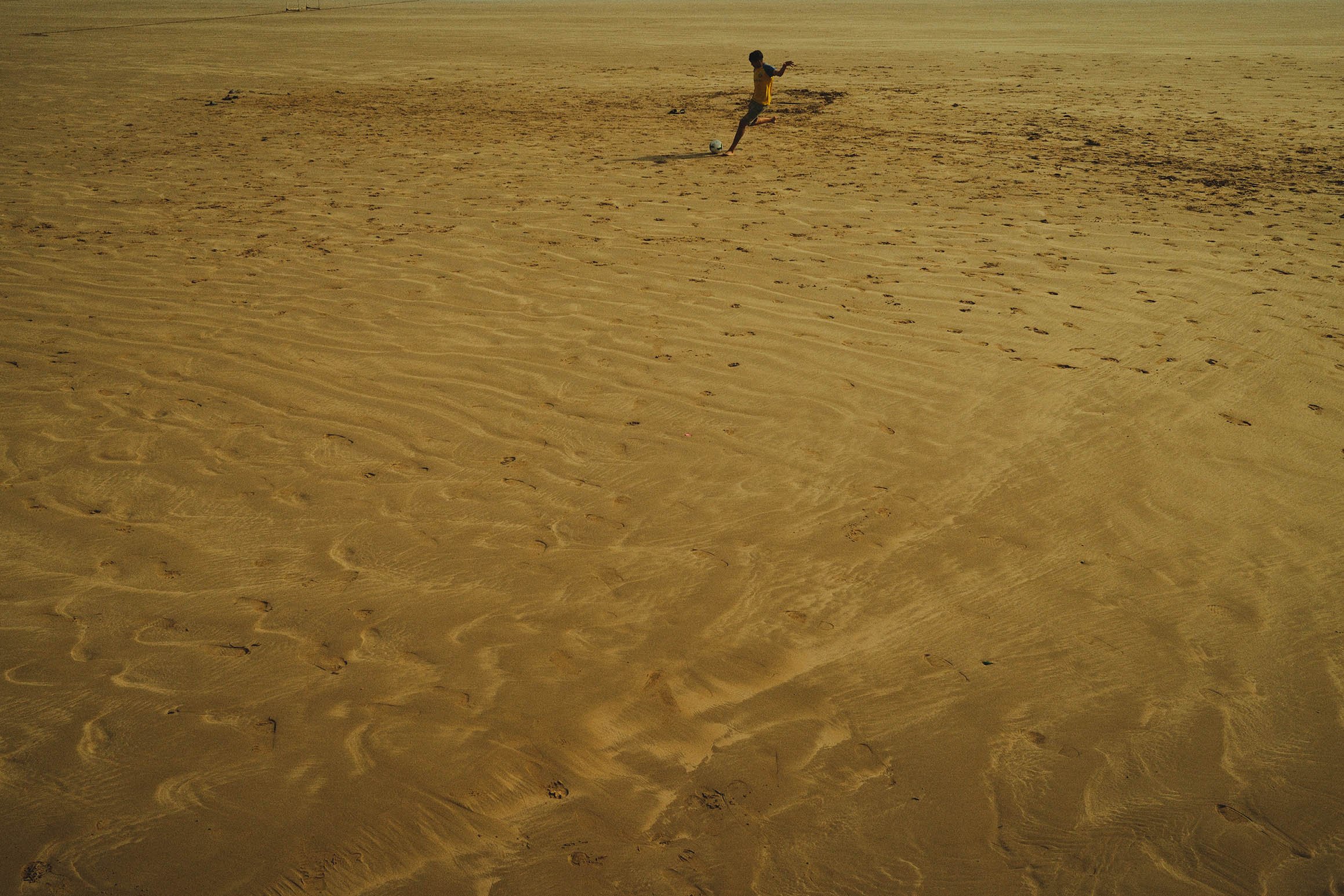 Un adolescent jouant au football sur une plage de sable. Le soleil brille et il n'y a pas d'autres personnes visibles.