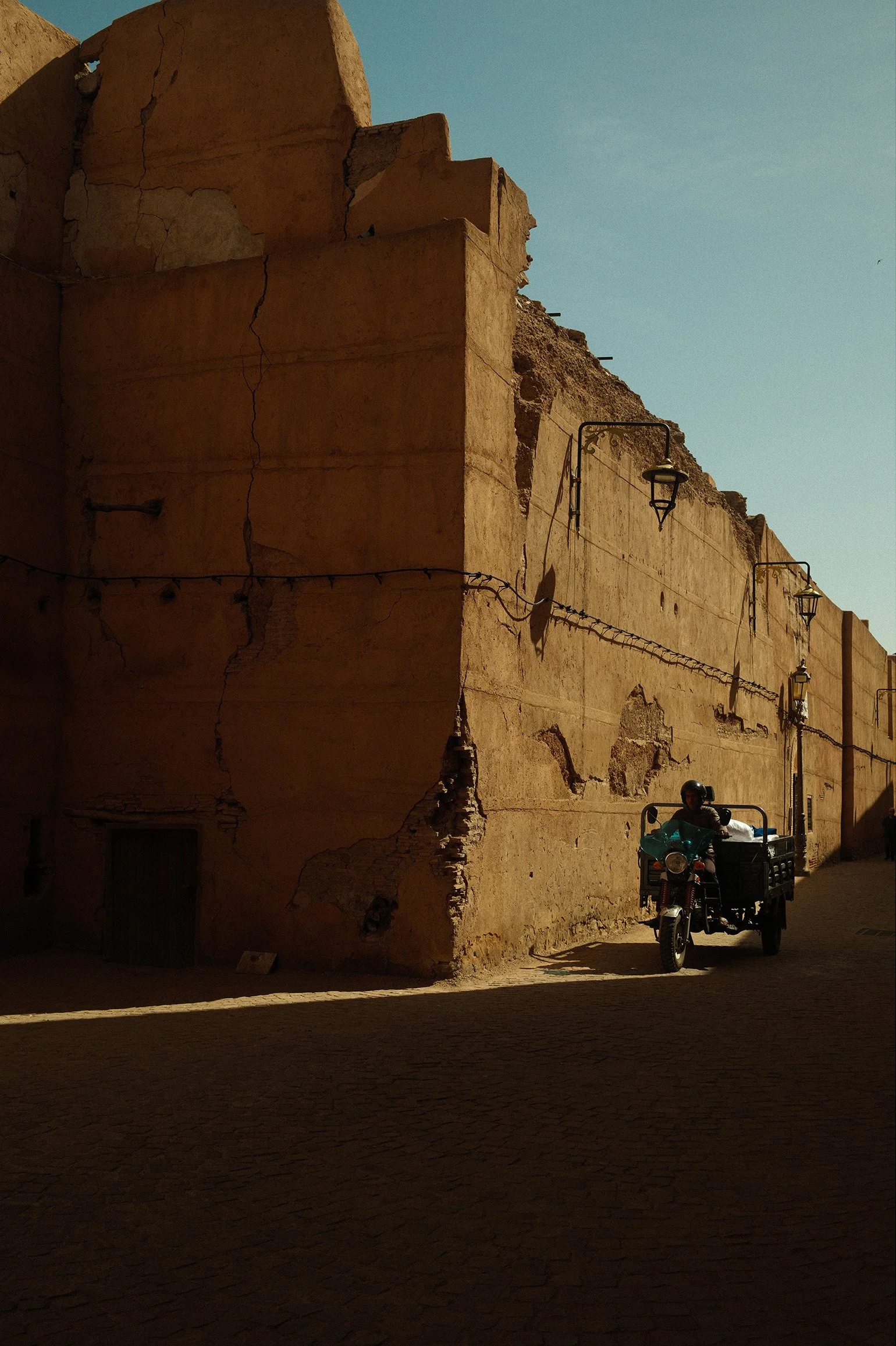 Une moto avec une personne portant un casque dans une rue bordée de murs en terre cuite endommagés, avec un ciel clair en arrière-plan.
