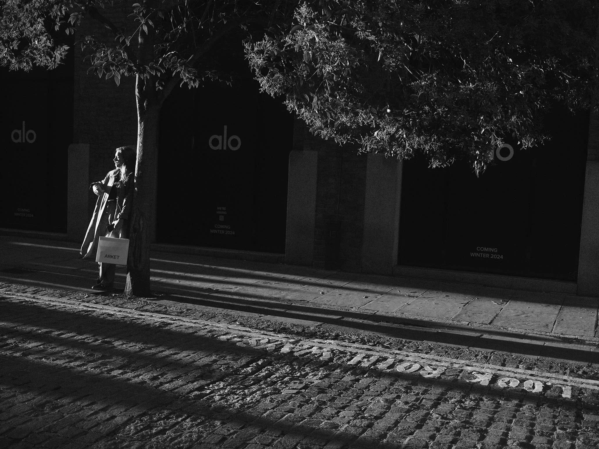 Une femme debout près d'un arbre, tenant un sac portant l'inscription 'ARKET', dans une rue pavée la nuit, avec des bâtiments en arrière-plan et des affiches annonçant la collection hiver 2024.