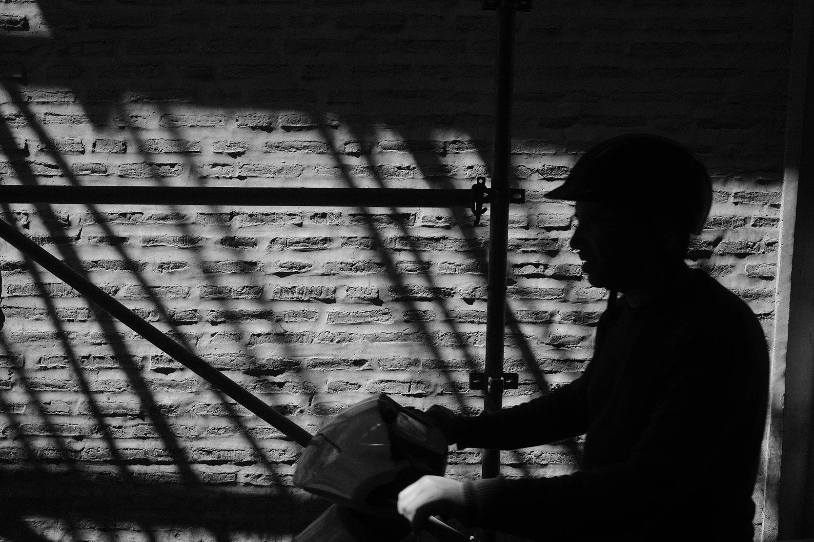 Silhouette d'un homme portant un casque, tenant un scooter, devant un mur en briques avec des ombres de structures métalliques.