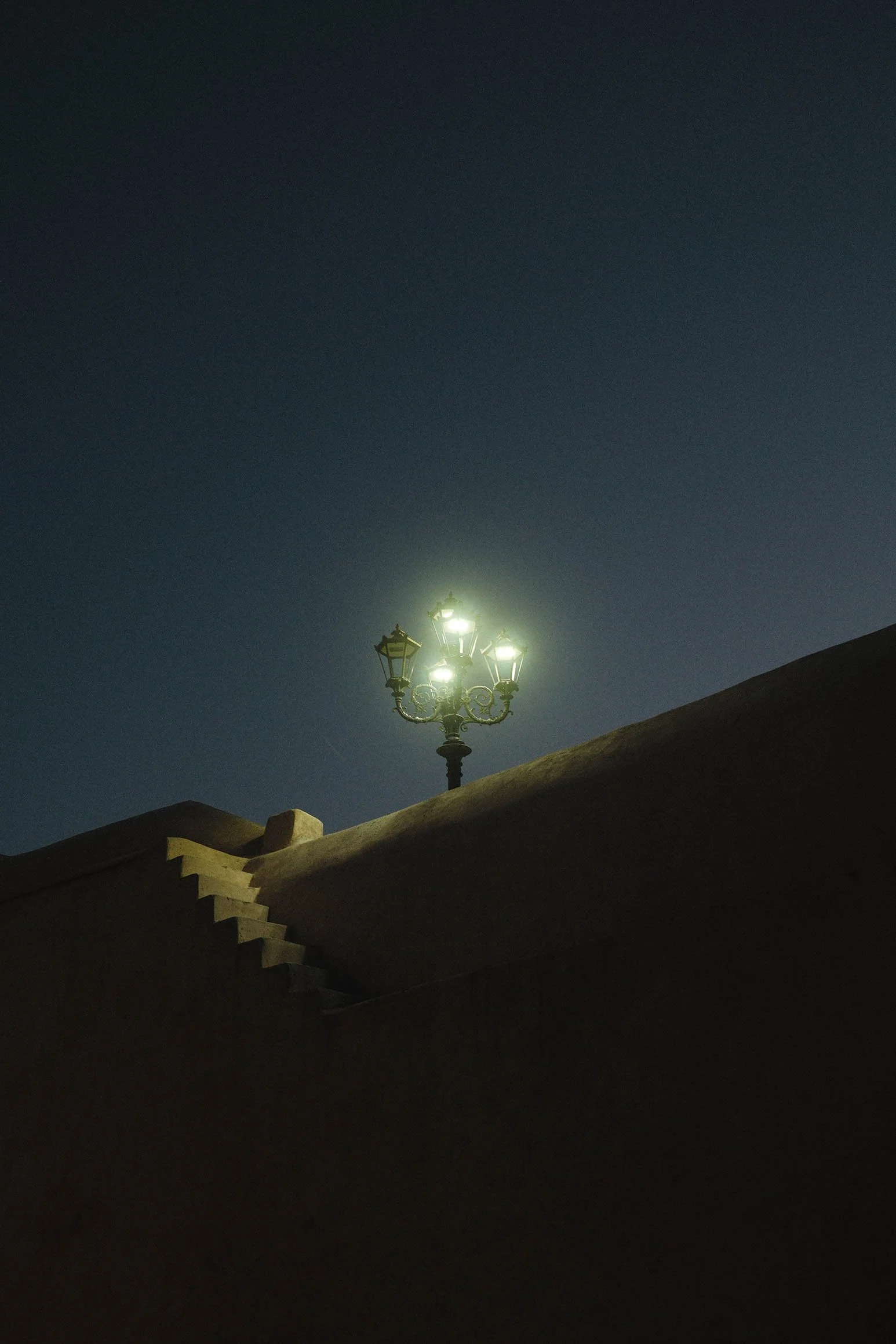 Un lampadaire éclairé la nuit, situé en haut d'une marche d'escalier en pierre, contre un ciel sombre.