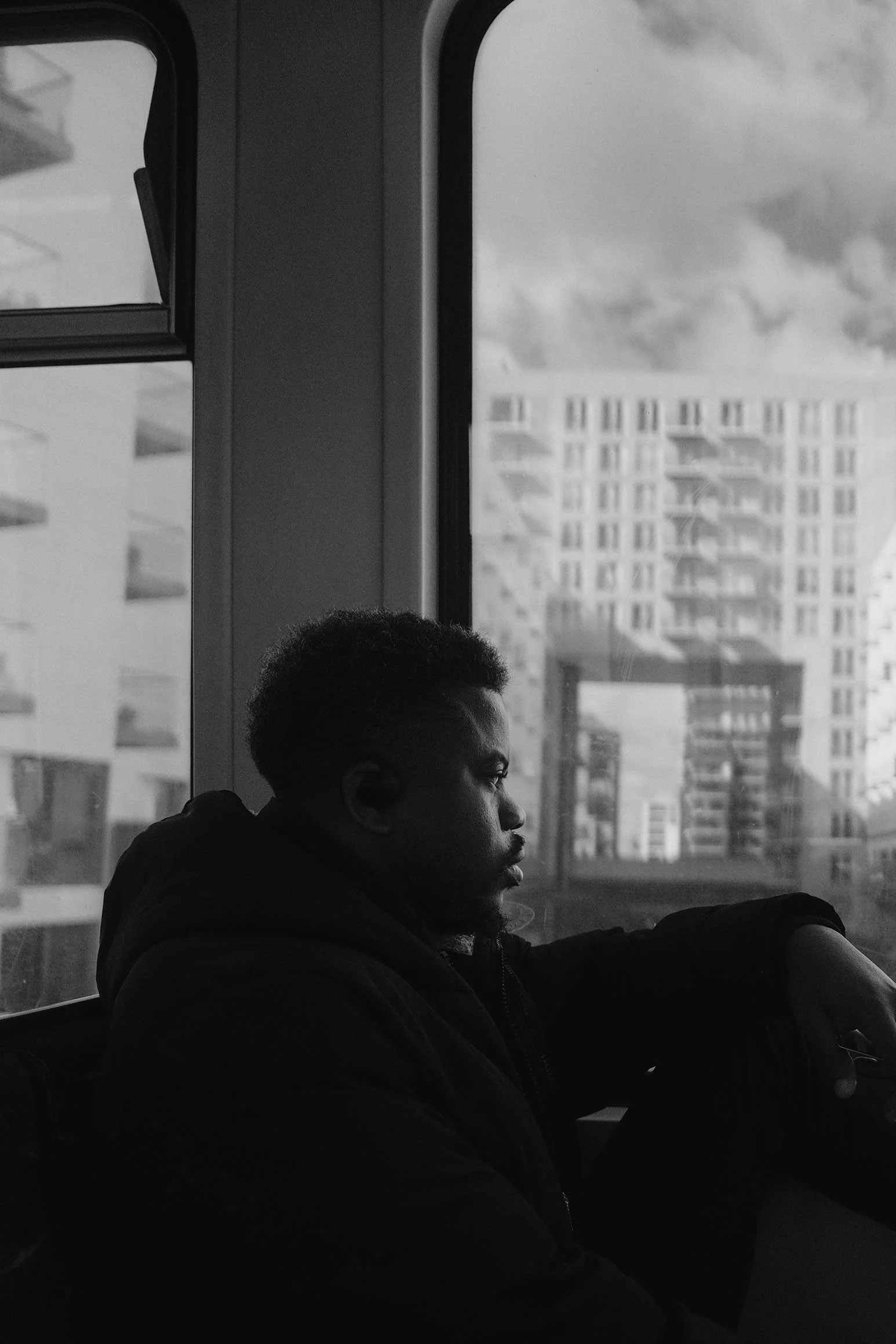 Un homme assis à une fenêtre dans un train, regardant dehors, en noir et blanc. La scène inclut des bâtiments modernes en arrière-plan et le ciel nuageux.