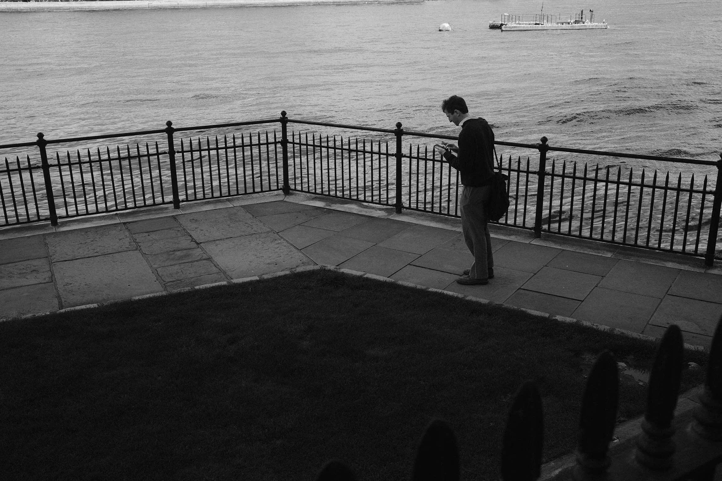 Un homme se tient sur un quai en pierre près de la mer, regardant son téléphone portable. Il porte un sac à l'épaule et porte une veste noire. La mer en arrière-plan est calme, avec un bateau au loin.