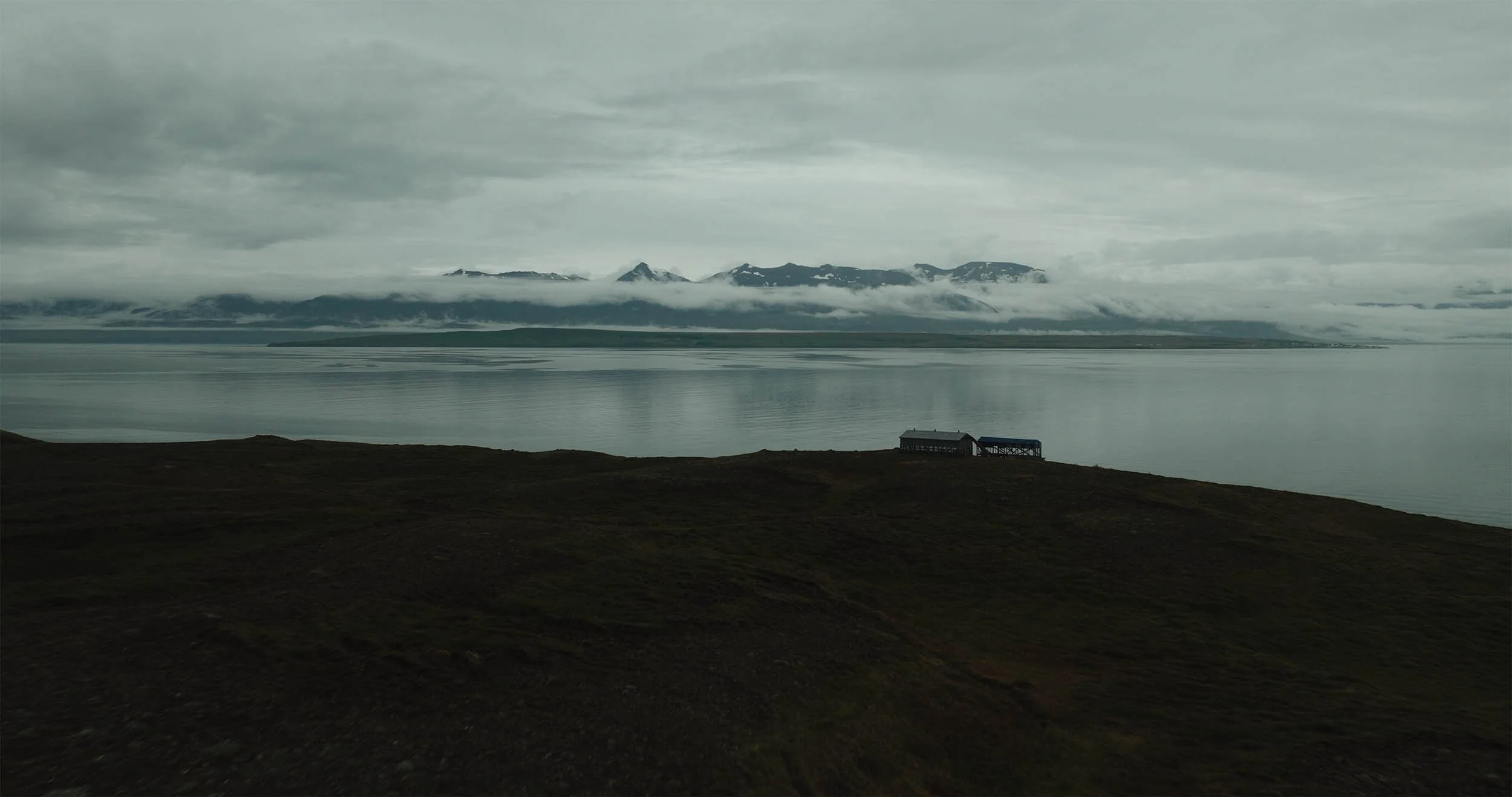 Paysage avec une mer calme, des montagnes en arrière-plan avec des sommets enneigés et une cabane en bois sur la rive en premier plan.