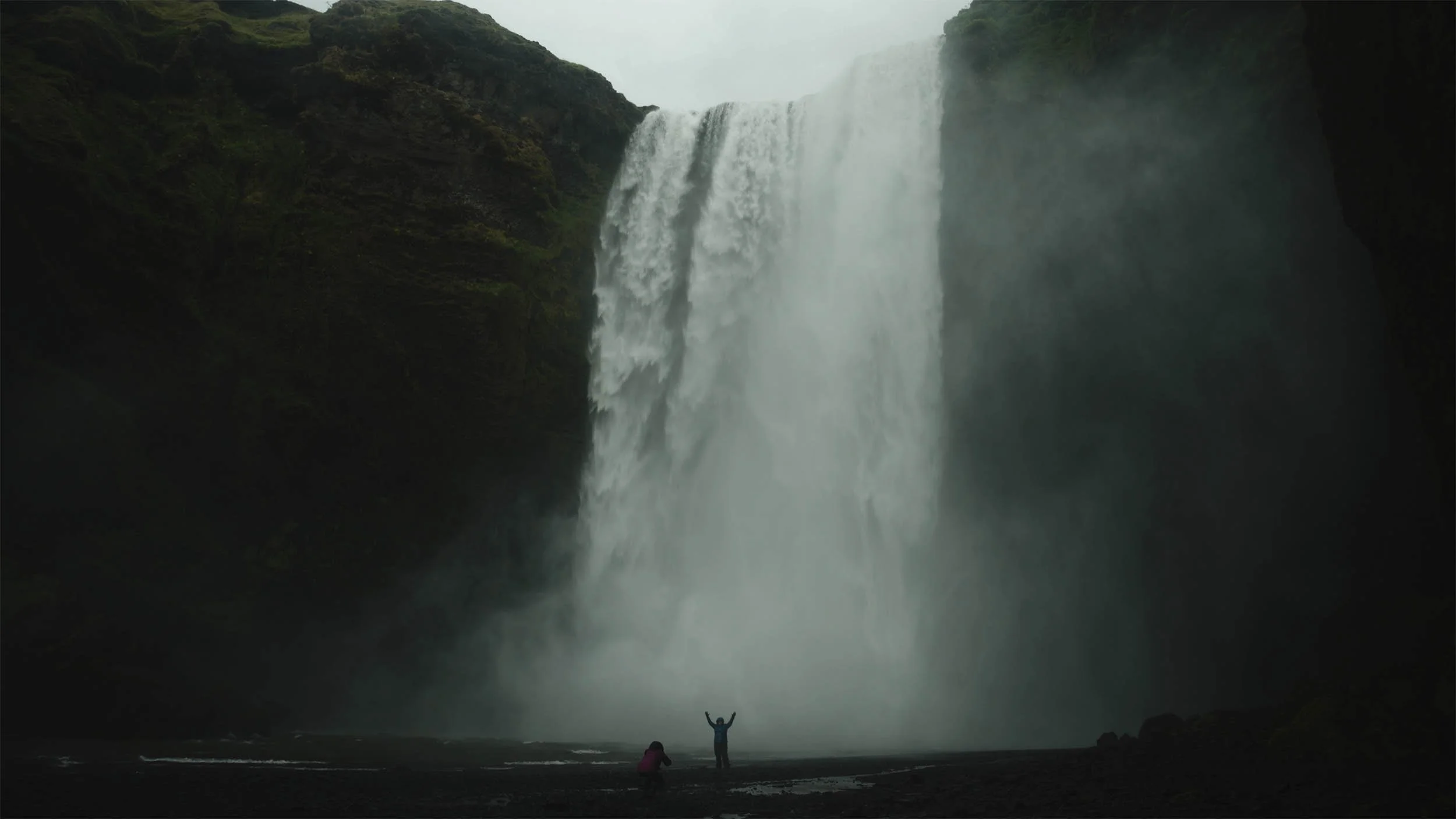 Une cascade impressionnante avec deux personnes au premier plan, une levant les bras et l'autre prenant une photo, entourée d'une nature verdoyante et sombre.