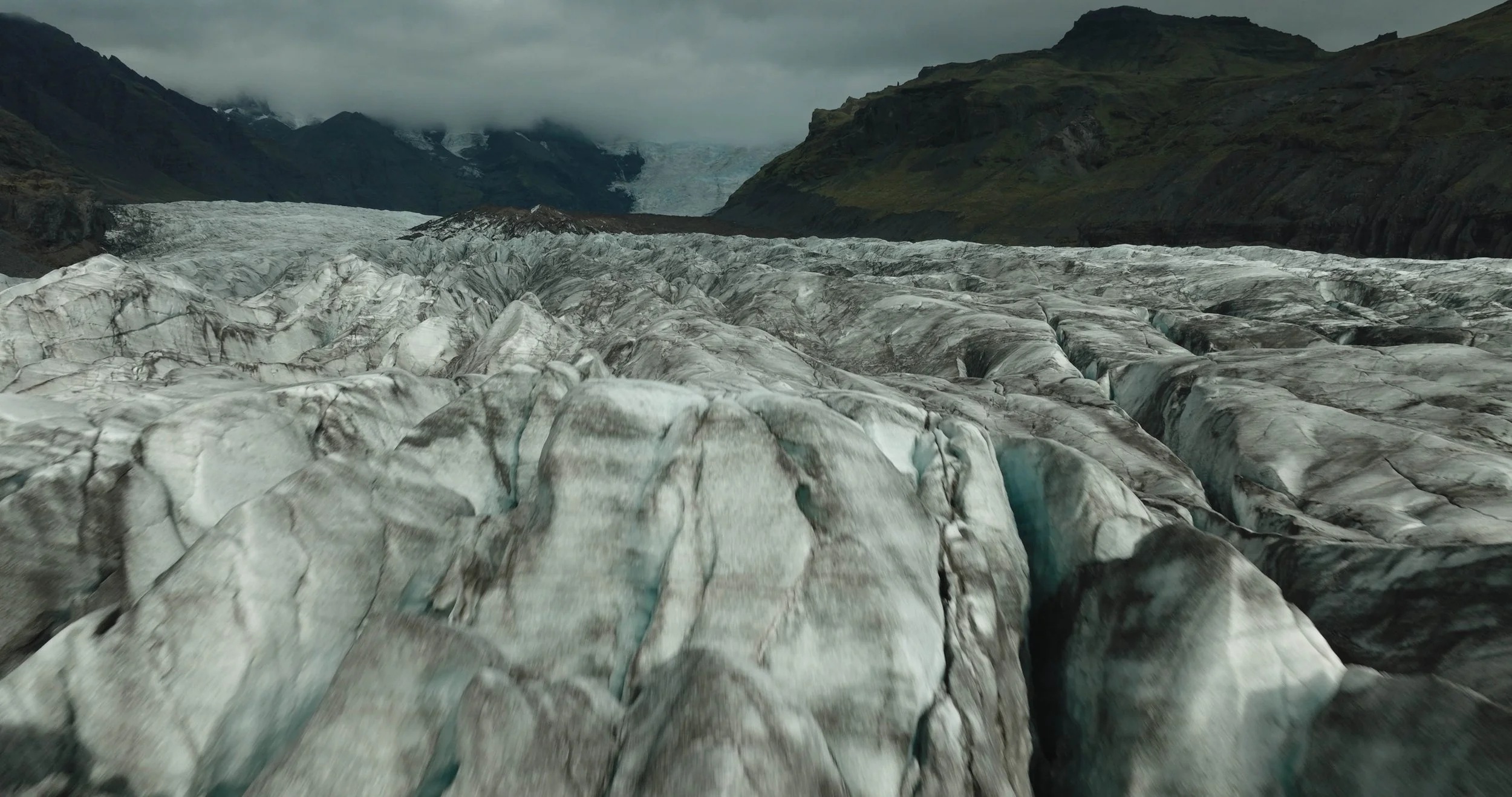 Un glacier avec des crevasses et des montagnes en arrière-plan sous un ciel nuageux.