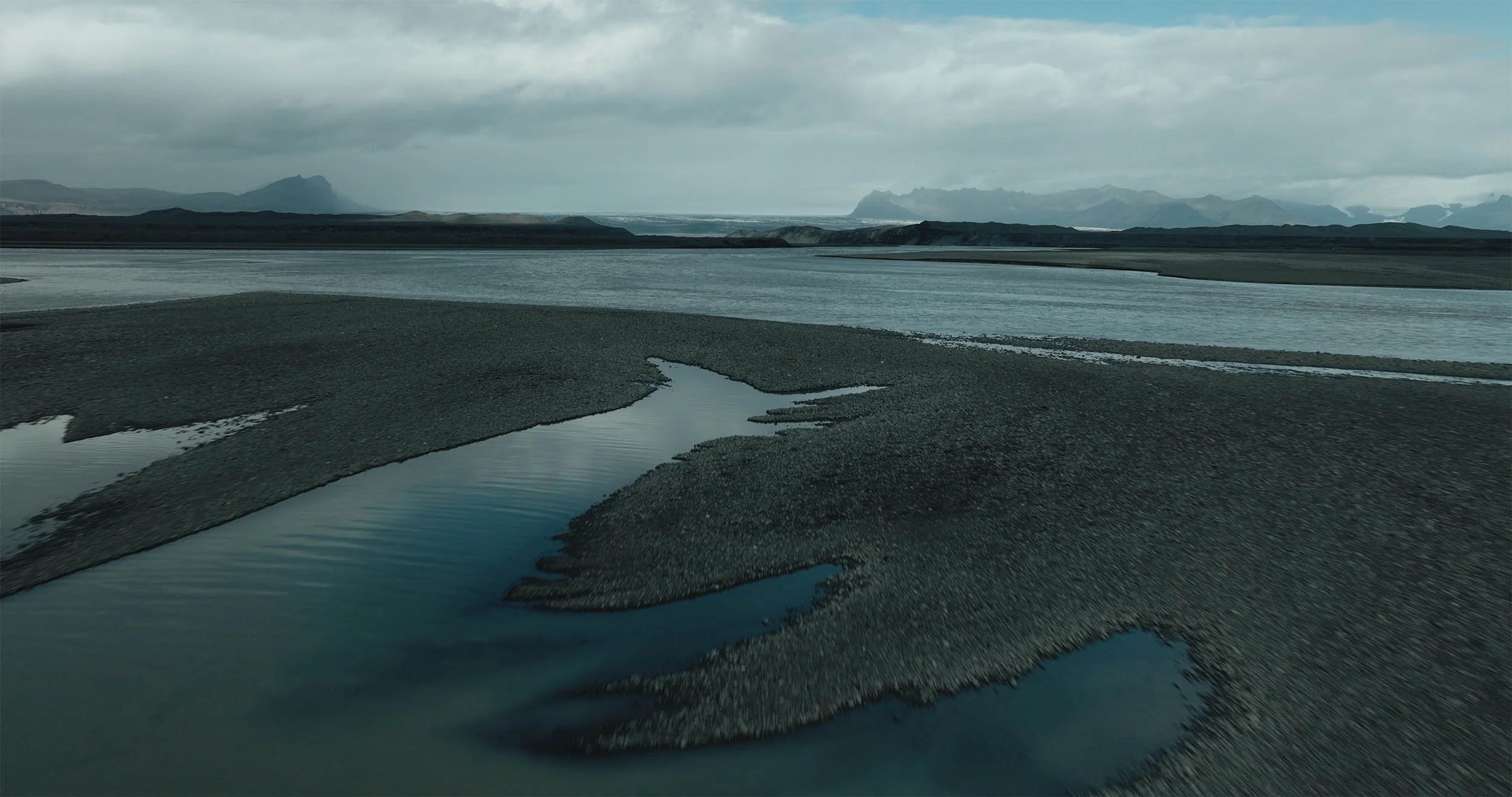 Paysage avec un fleuve serpentant à travers un terrain rocheux et plat, montagnes à l'arrière-plan, ciel nuageux.