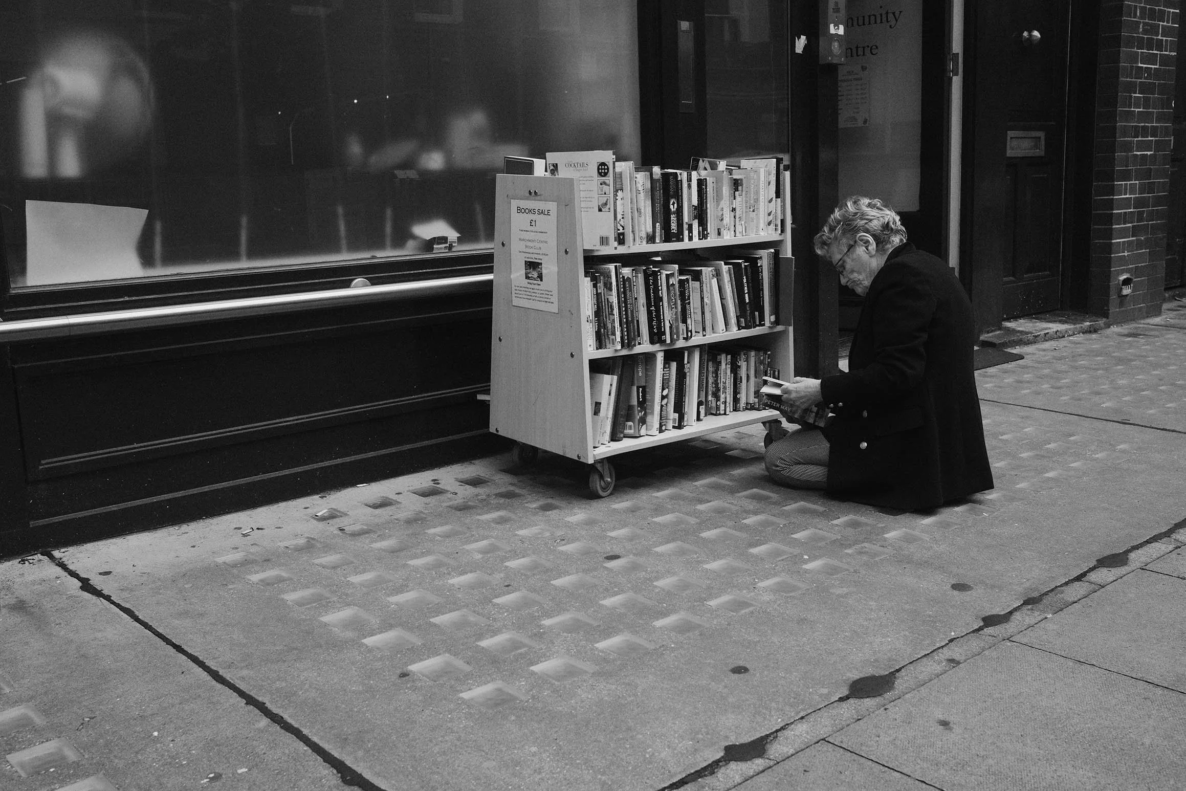 Une femme âgée à genoux devant une étagère à livres dehors, lisant un livre. Il y a une affiche pour une vente de livres à 1 euro attachée à l'étagère.