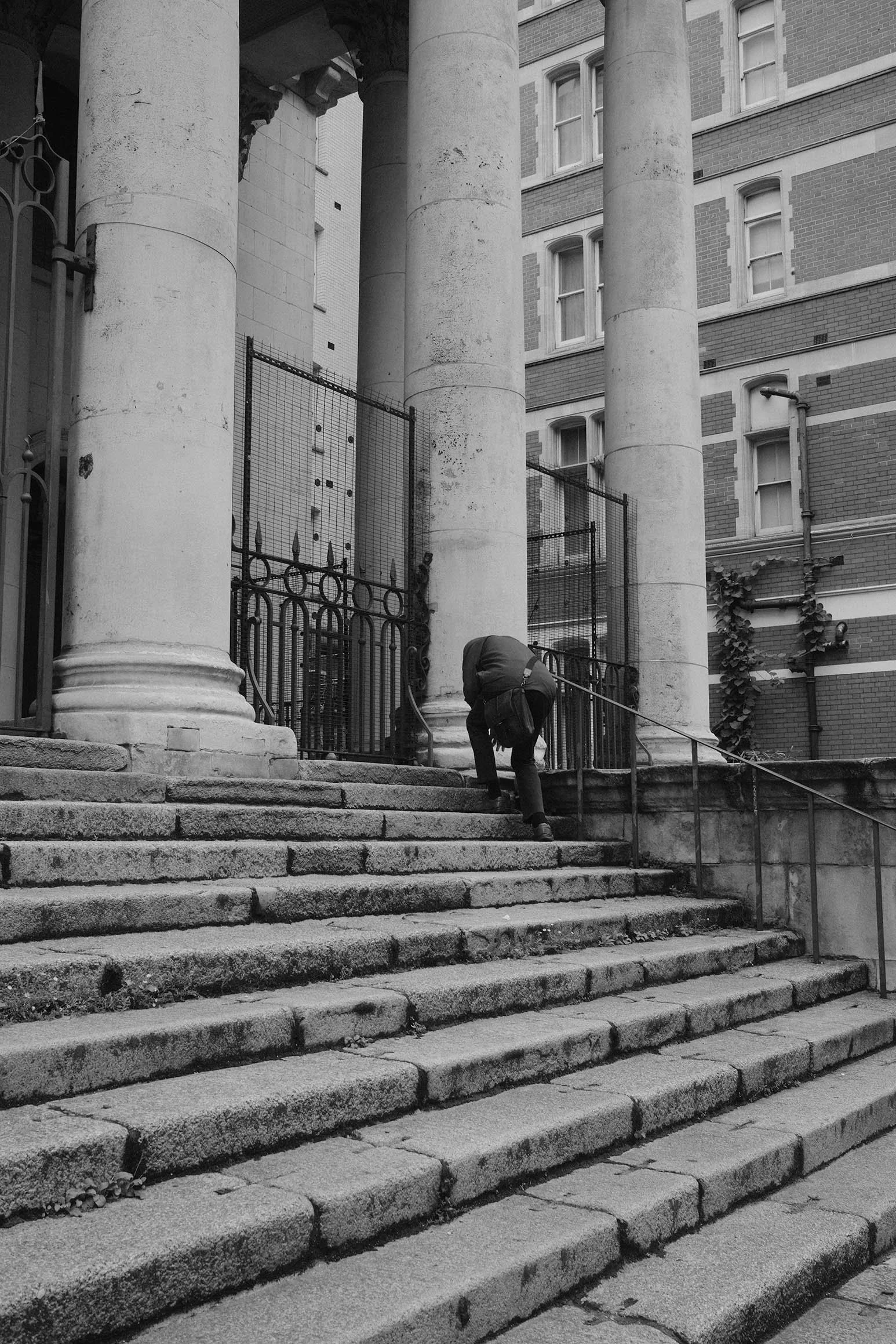 Un homme grimpe les escaliers en pierre menant à un bâtiment avec de grandes colonnes et un immeuble en briques en arrière-plan.