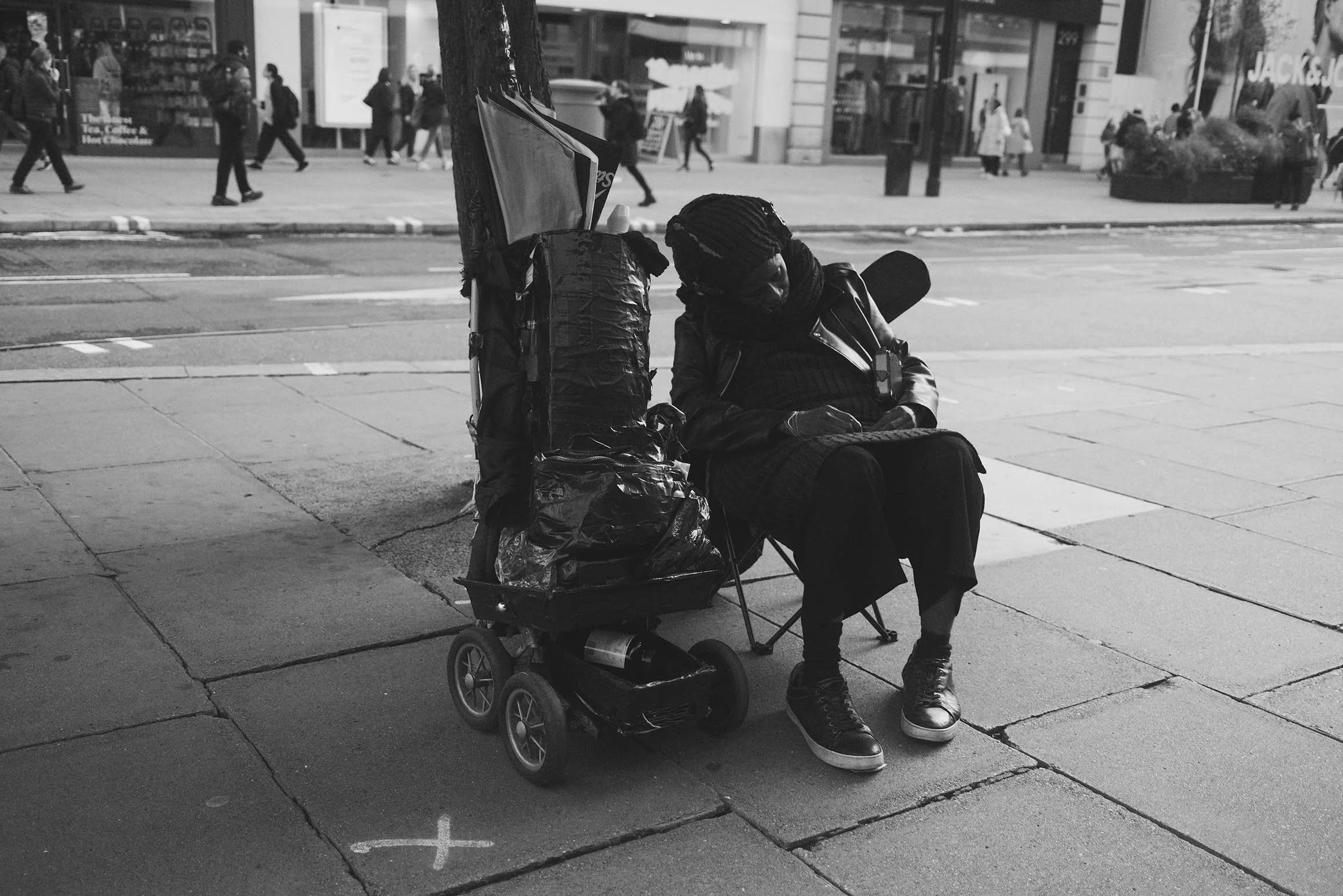 Une femme sans domicile fixe assise sur le trottoir avec un chariot chargé de sacs et d'articles, en ville