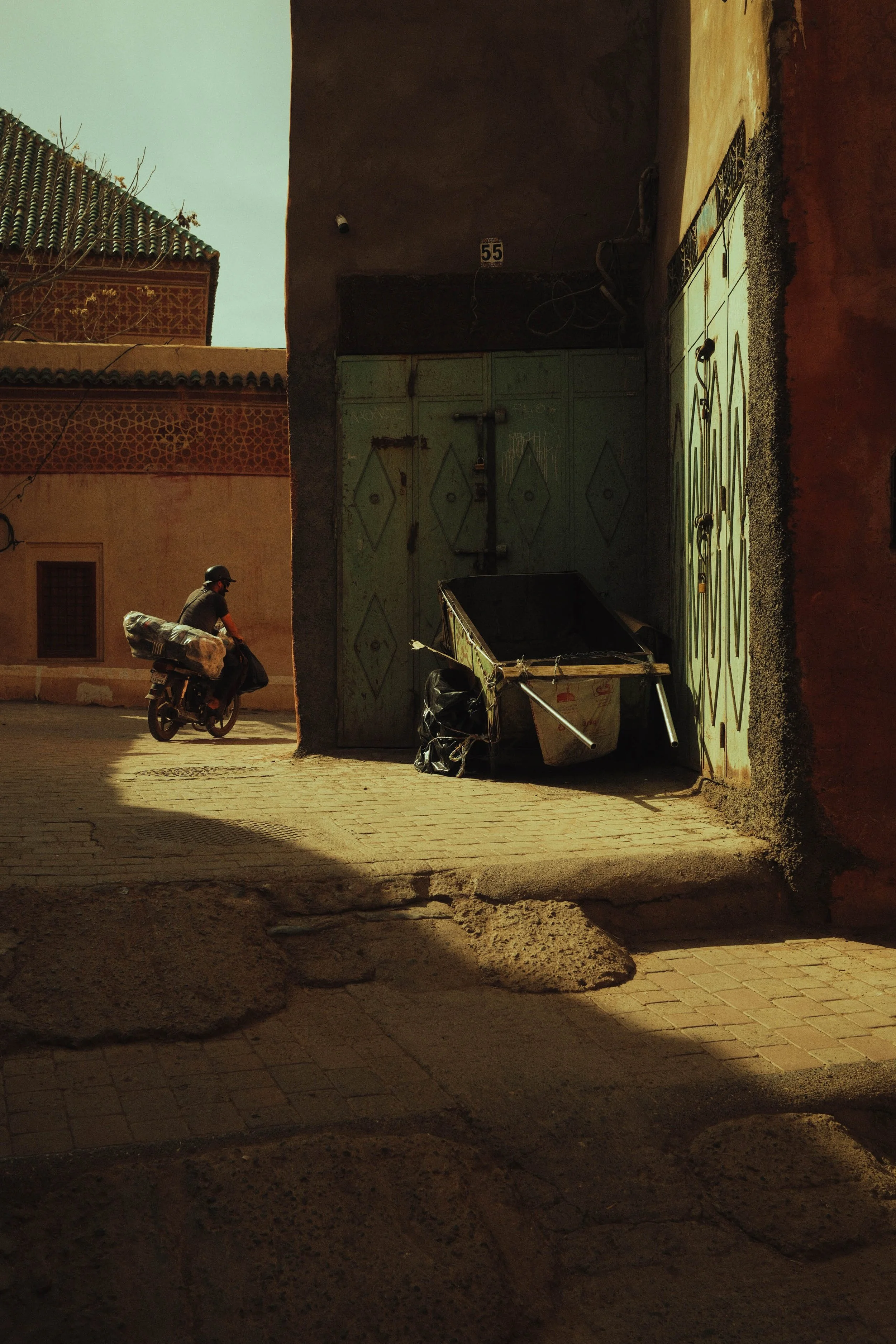 Un homme à vélo passe devant un mur ensoleillé avec une grande porte métallique et un caddy de transport abandonné. La scène est dans une zone urbaine, avec des murs de couleur rouge et une ombre portée sur le sol pavé.