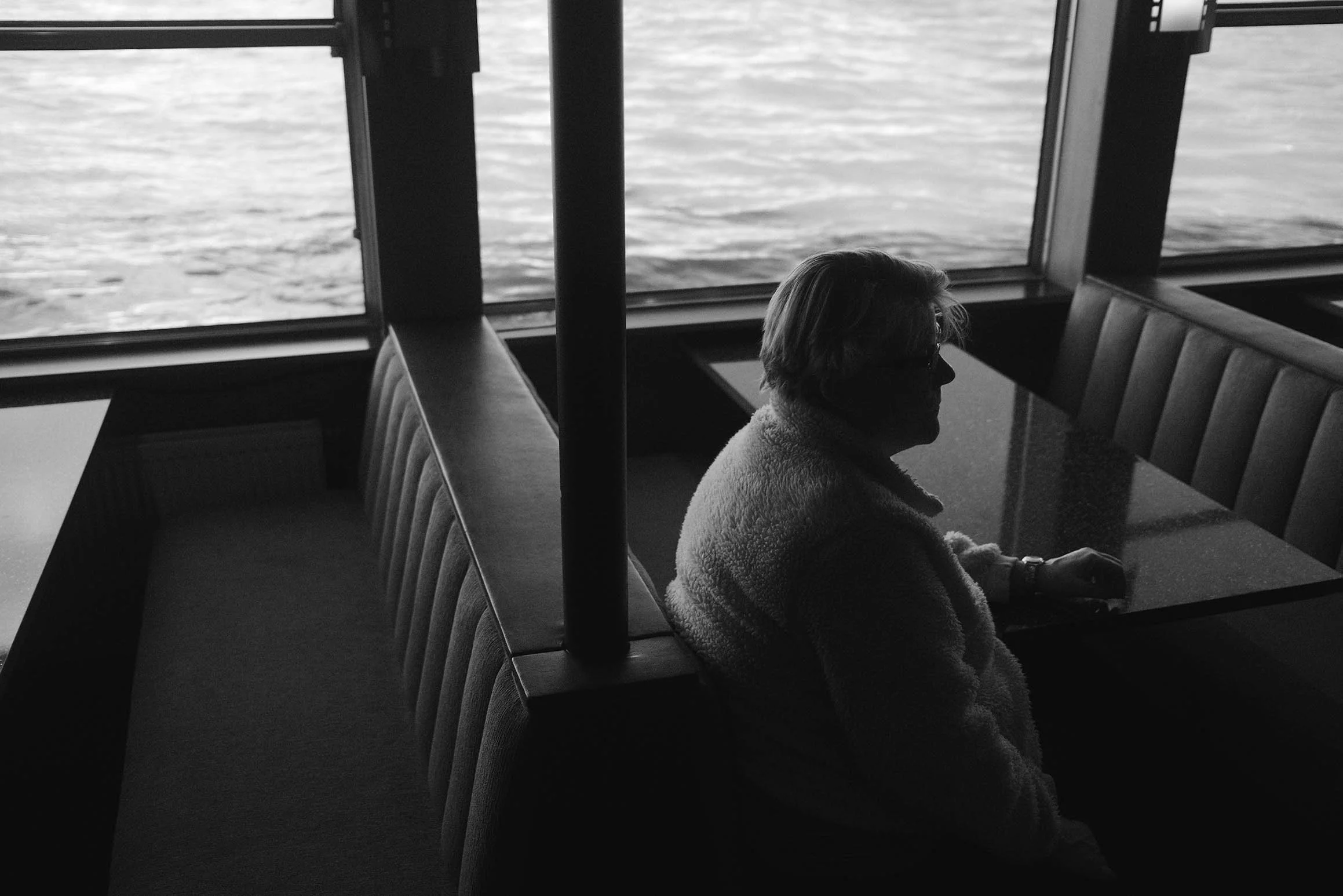 Une personne âgée assise dans un coin d'un restaurant face à une grande fenêtre donnant sur un plan d'eau, vue en noir et blanc.
