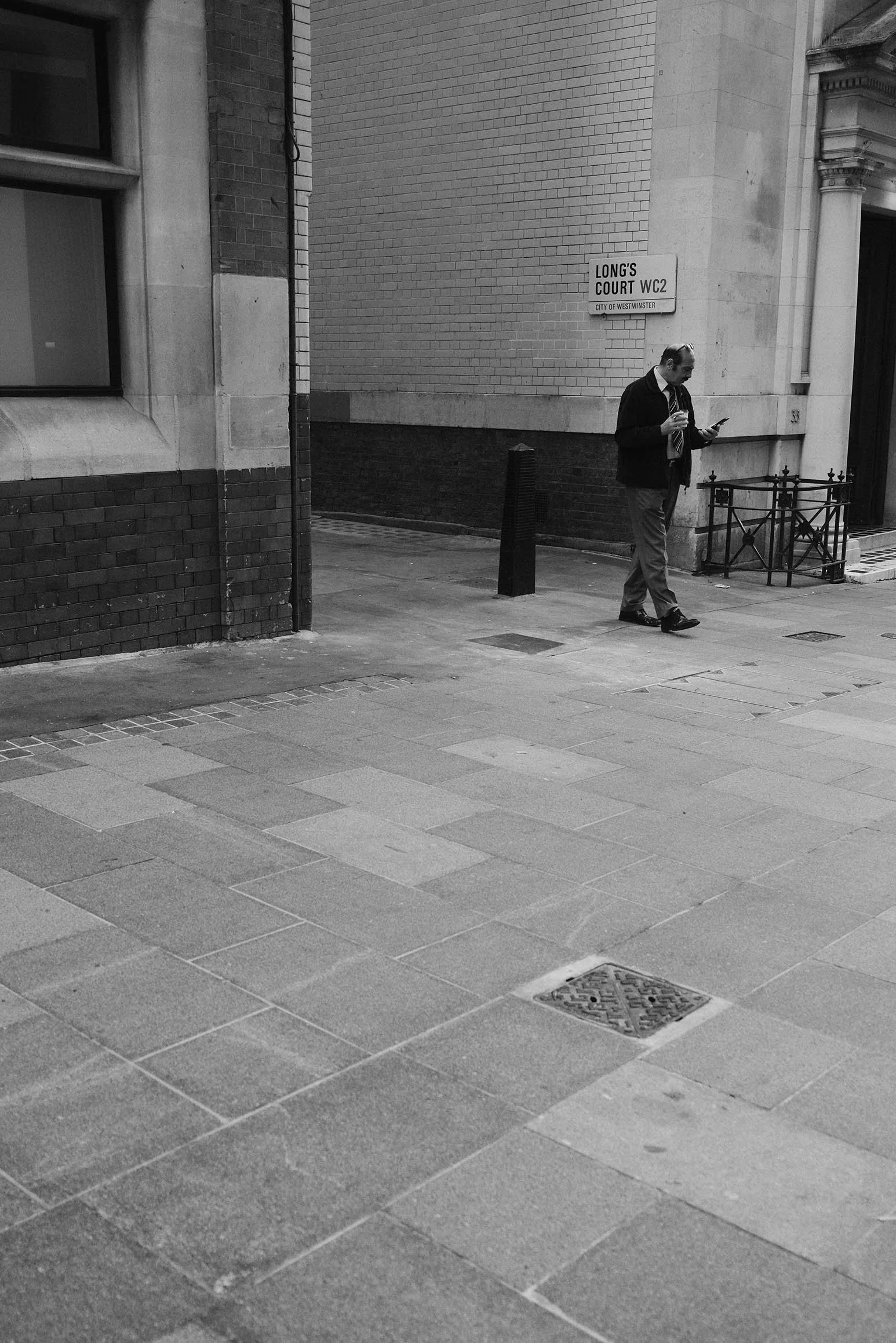 Un homme d'affaires marchant dans la rue en regardant son téléphone, devant un bâtiment avec un panneau indiquant 'Long's Court WC2', dans une scène en noir et blanc.
