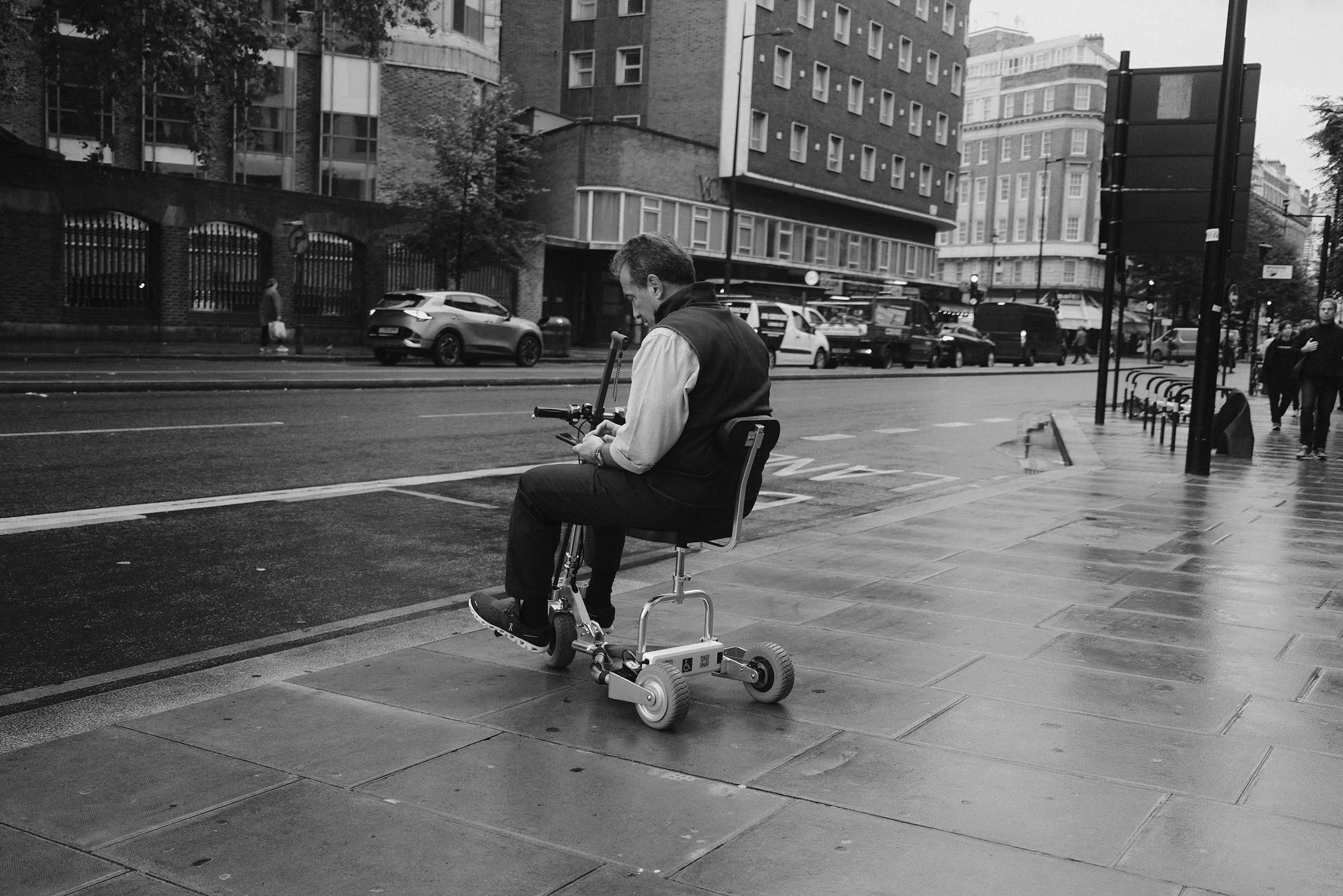 Un homme assis sur un fauteuil roulant électrique sur un trottoir d'une rue urbaine, regardant son téléphone, en noir et blanc.