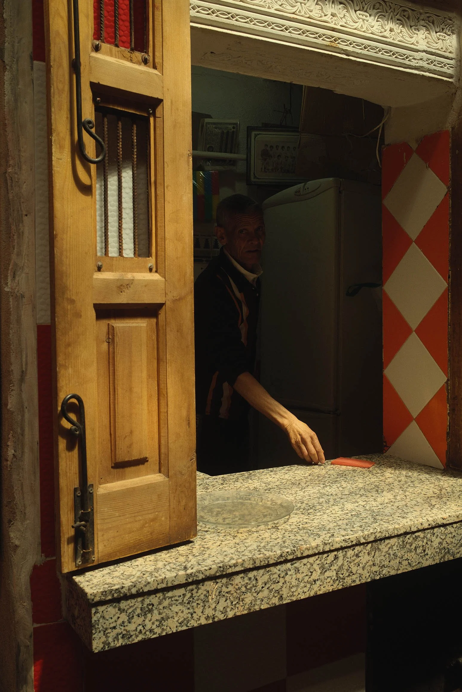 Un homme regarde à travers une fenêtre avec un cadre en bois, tenant une plaque rouge-orange sur un comptoir en granite, dans une cuisine avec un réfrigérateur et une décoration murale en relief.