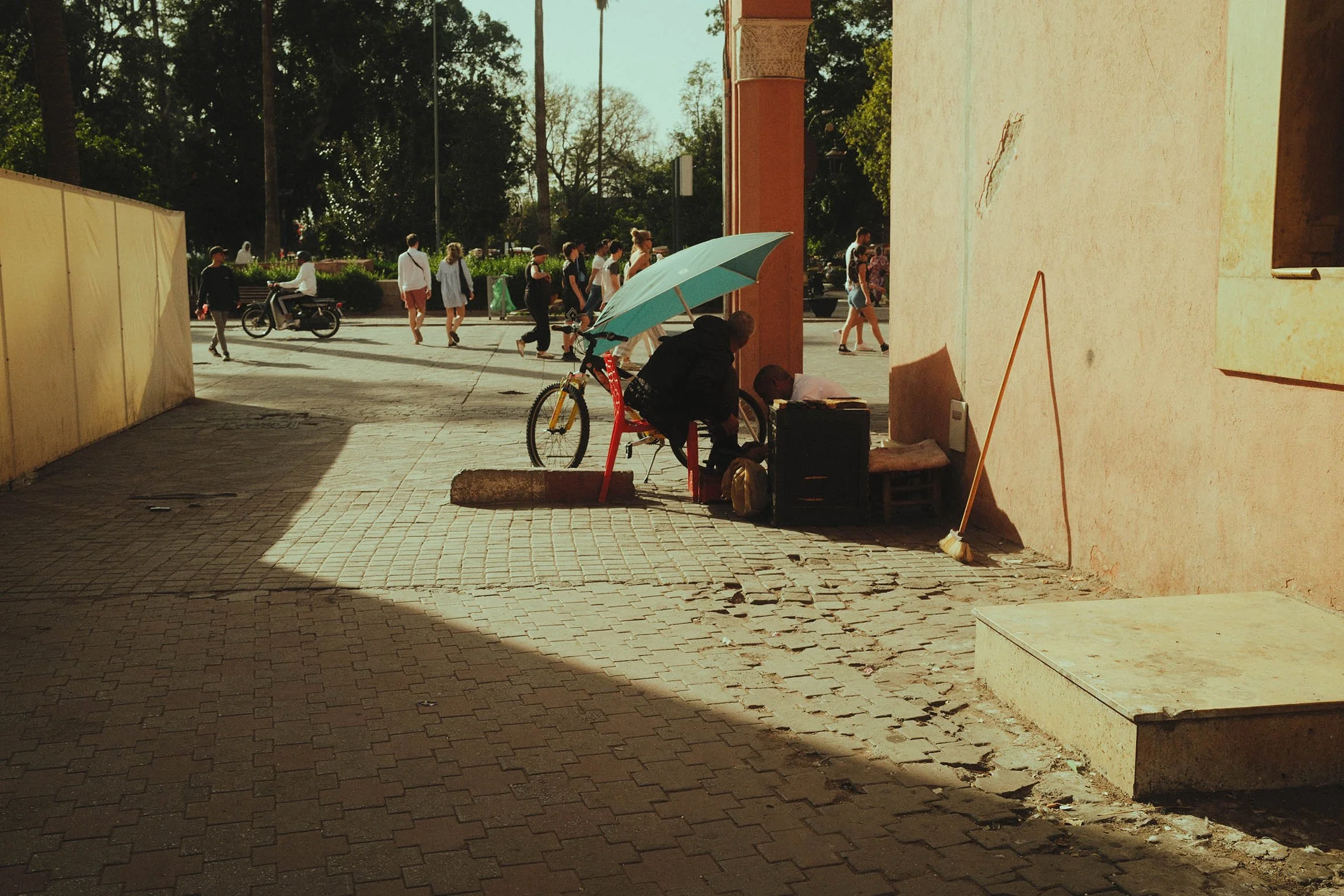 Personne sous un parasol bleu, assise à un comptoir, près d'une bicyclette, dans une rue animée, avec des passants et des arbres en arrière-plan.