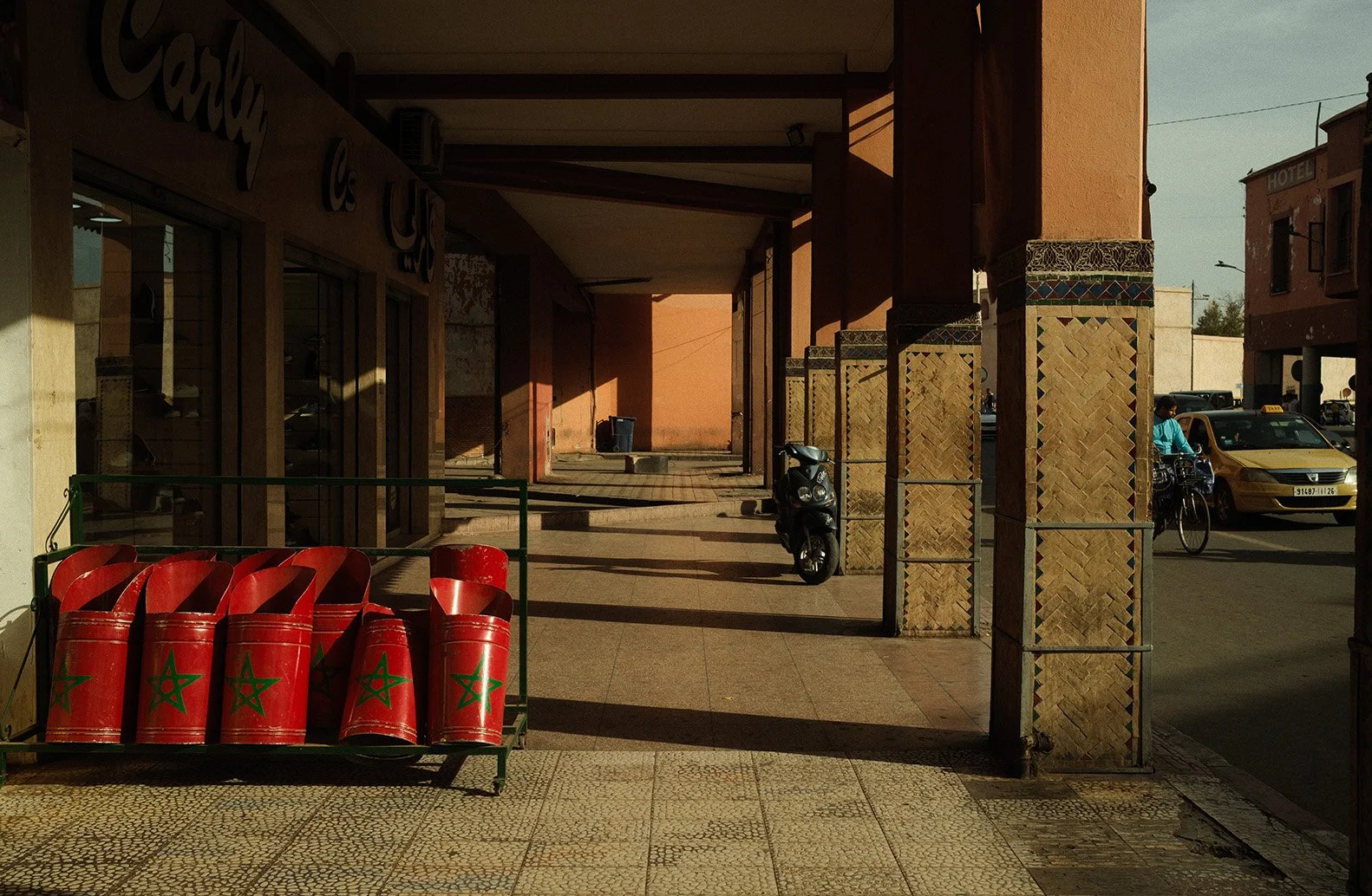 Vitrine d'une boutique avec des poubelles rouges portant une étoile verte, un scooter stationné devant un bâtiment, et une personne à vélo passant devant des voitures jaunes, dans un environnement urbain ensoleillé.