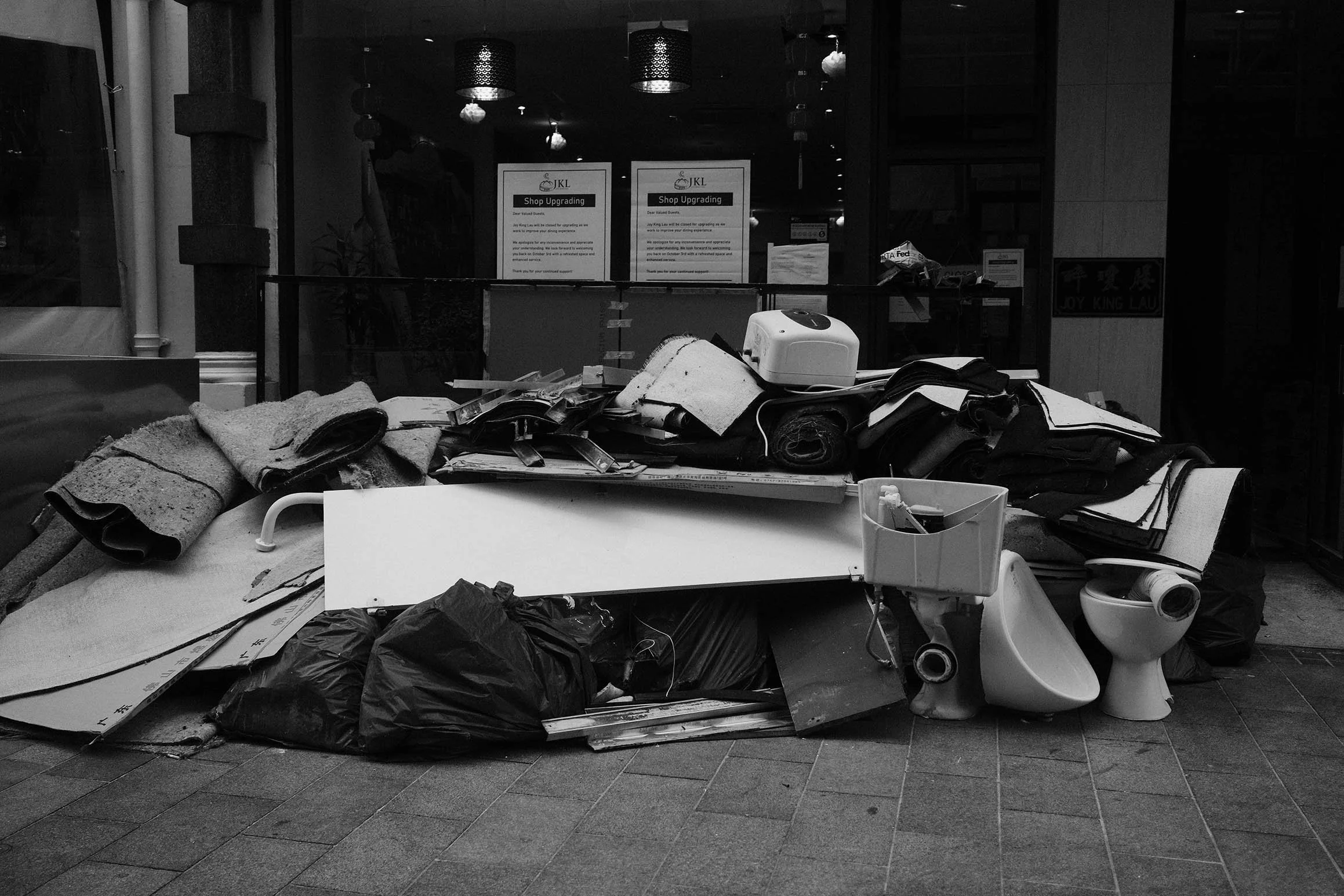 Une montagne de débris et d'objets abandonnés, comprenant des meubles endommagés, des sacs en plastique, et du matériel divers, devant une vitrine d'un magasin fermée.
